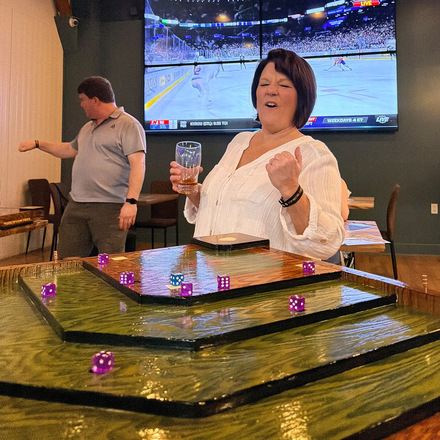 A player or group of friends are playing the dice-flicking party game, Baseball Shuffle, with drinks on the handcrafted oak game table. They are having a fun time at a brewery, taproom or social venue.