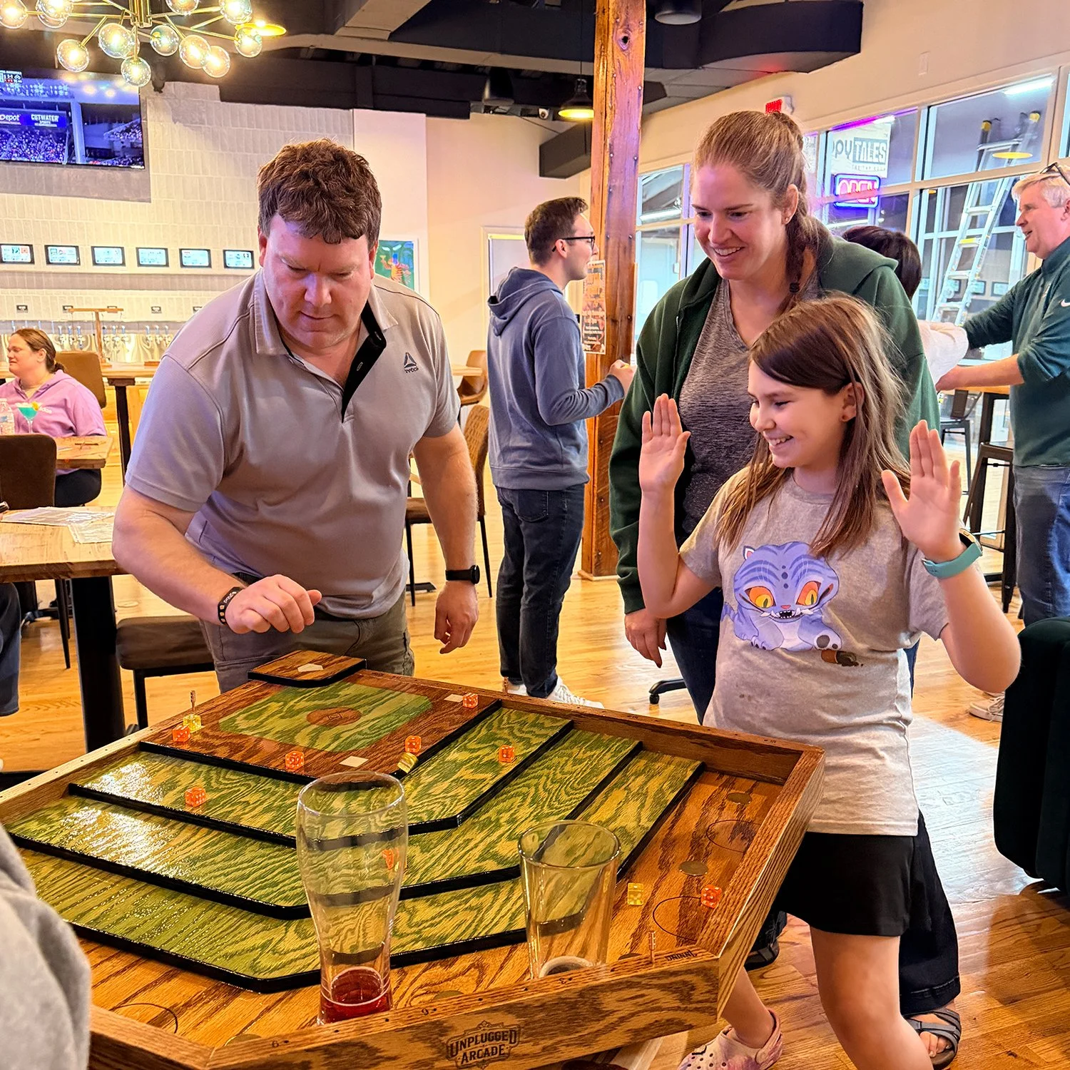 A player or group of friends are playing the dice-flicking party game, Baseball Shuffle, with drinks on the handcrafted oak game table. They are having a fun time at a brewery, taproom or social venue.