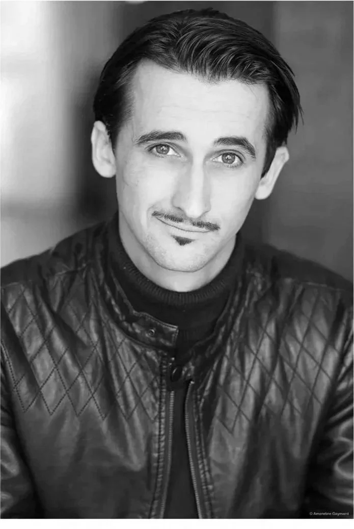 A man with dark hair, a mustache, and goatee, wearing a black quilted leather jacket, looking at the camera with a slight smile.