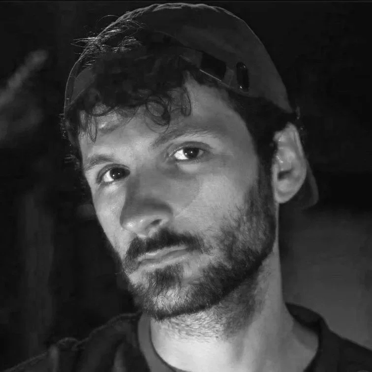 A young man with a beard and short dark hair, wearing a backwards cap, illuminated with colorful pink and blue lighting.