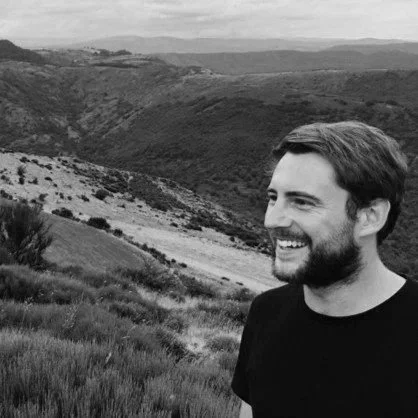 A man with brown hair and a beard smiling outdoors with green hills and mountains in the background.