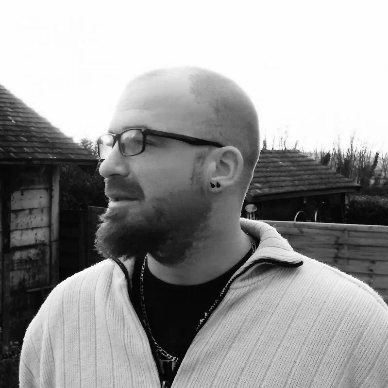 A man with glasses, a beard, and a side-shaved hairstyle stands outdoors in front of a wooden fence and garden shed during sunset.