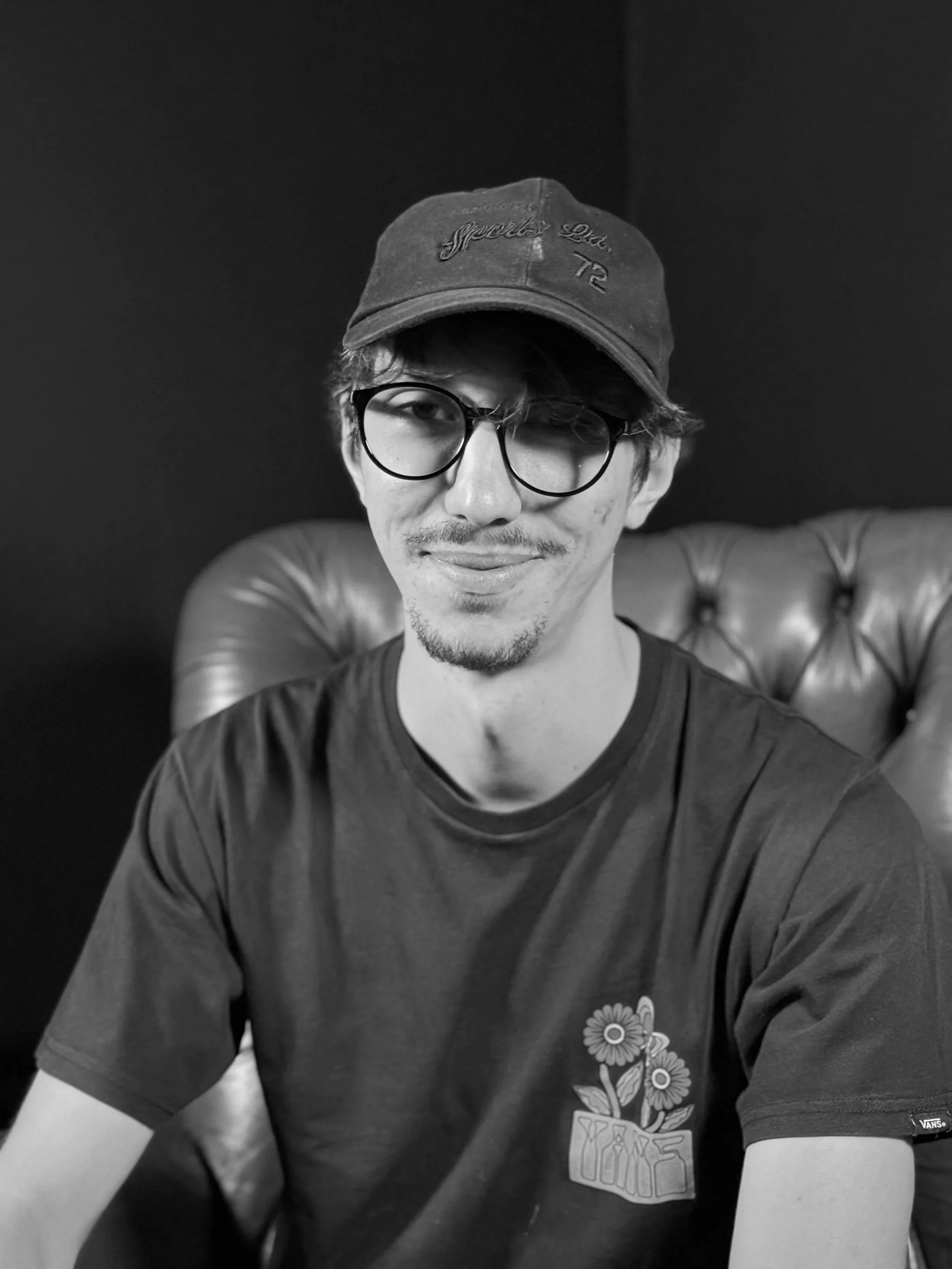 A young man with glasses and a goatee, wearing a dark t-shirt with a floral design, sitting in front of a dark background and a red leather armchair.