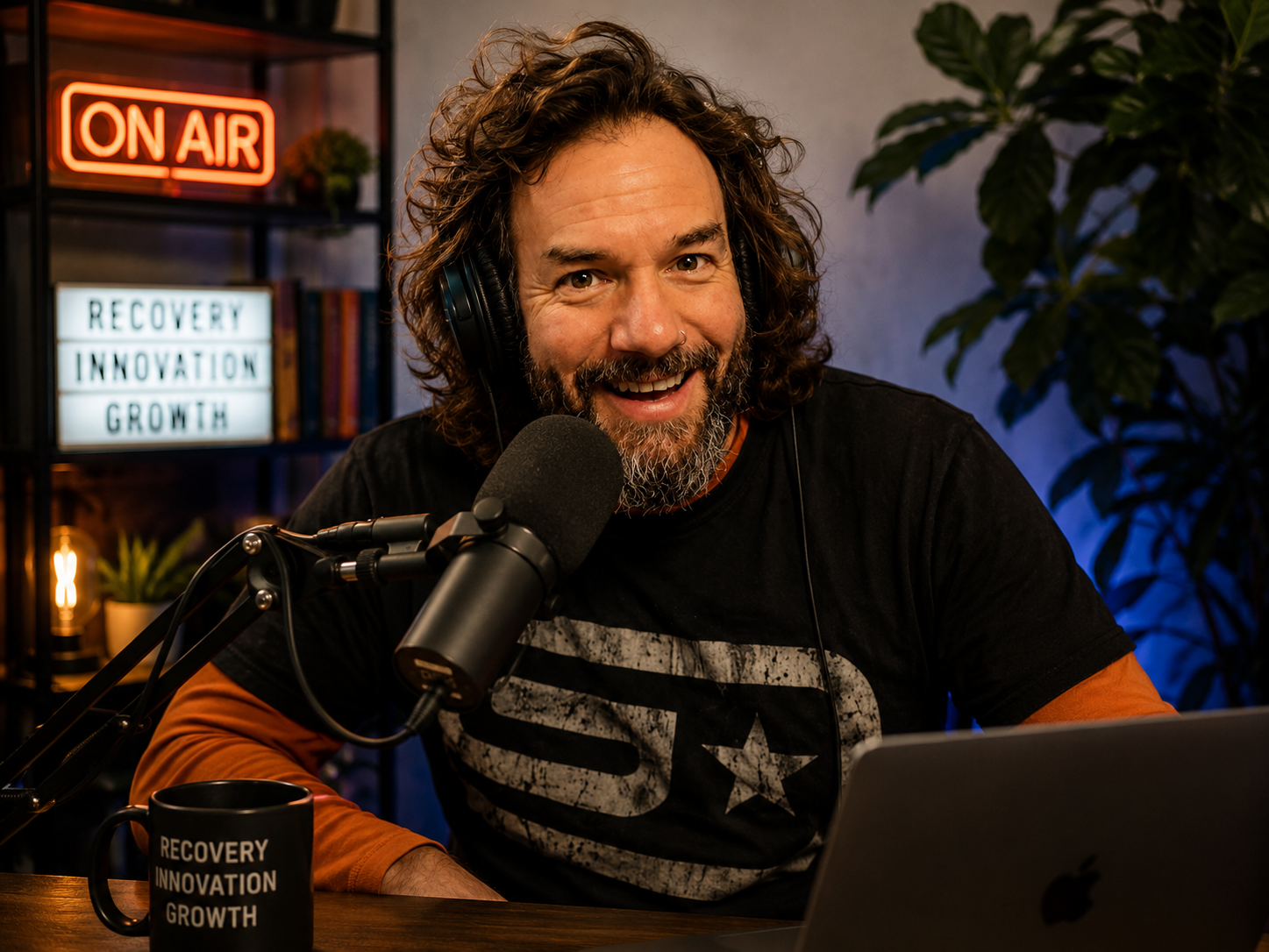 A man with curly hair and a beard wearing headphones, speaking into a microphone during a podcast recording session, with a laptop and a black mug with the words 'Recovery Innovation Growth' in front of him, in a studio with bookshelves, a neon 'On Air' sign, and indoor plants in the background.