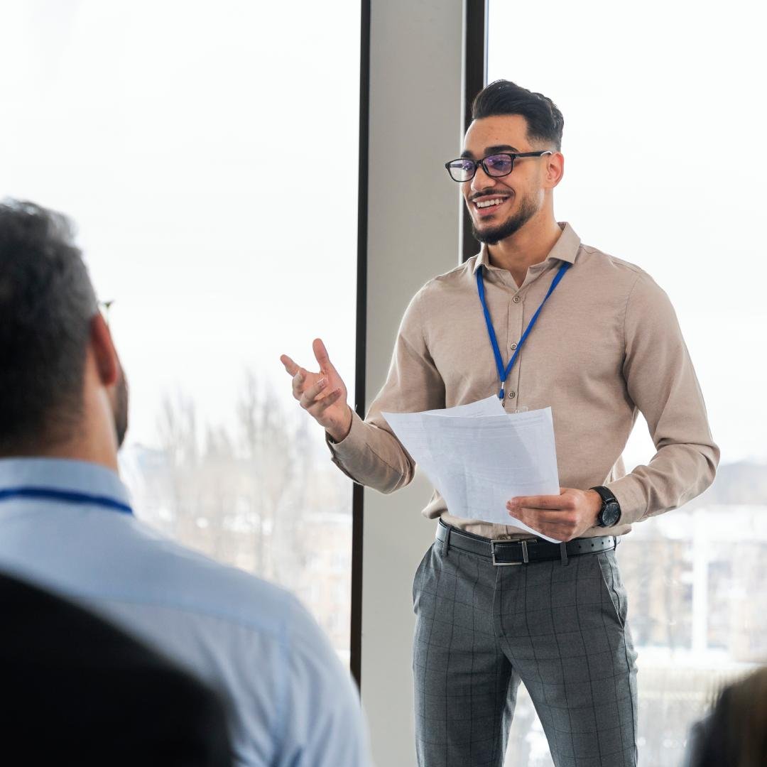 Homem apresentando em ambiente de escritório, segurando papéis, com dois colegas assistindo.