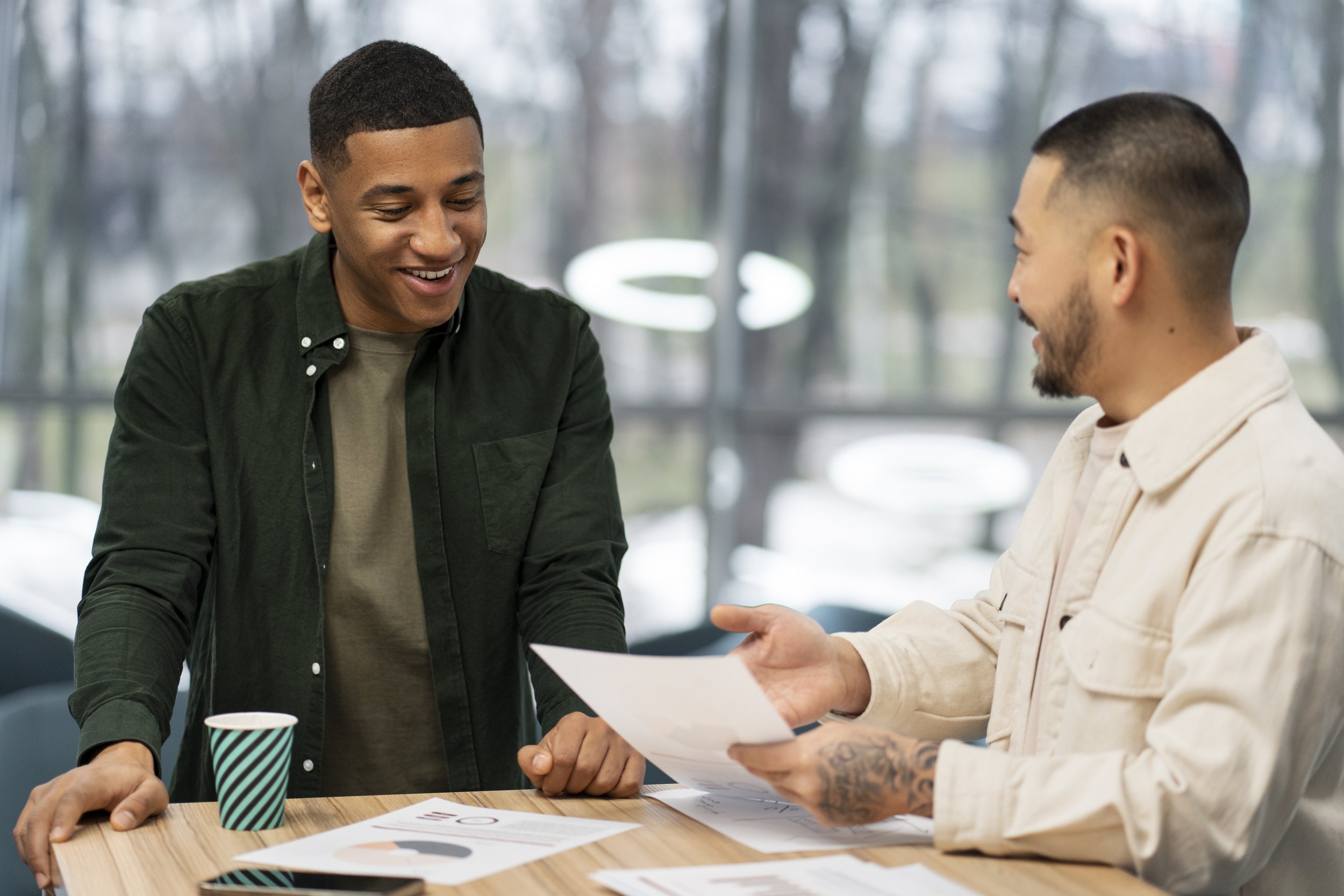 Duas pessoas sorrindo, uma transmitindo documentos a outra, em um ambiente de escritório com janelas grandes e vista de árvores ao fundo.