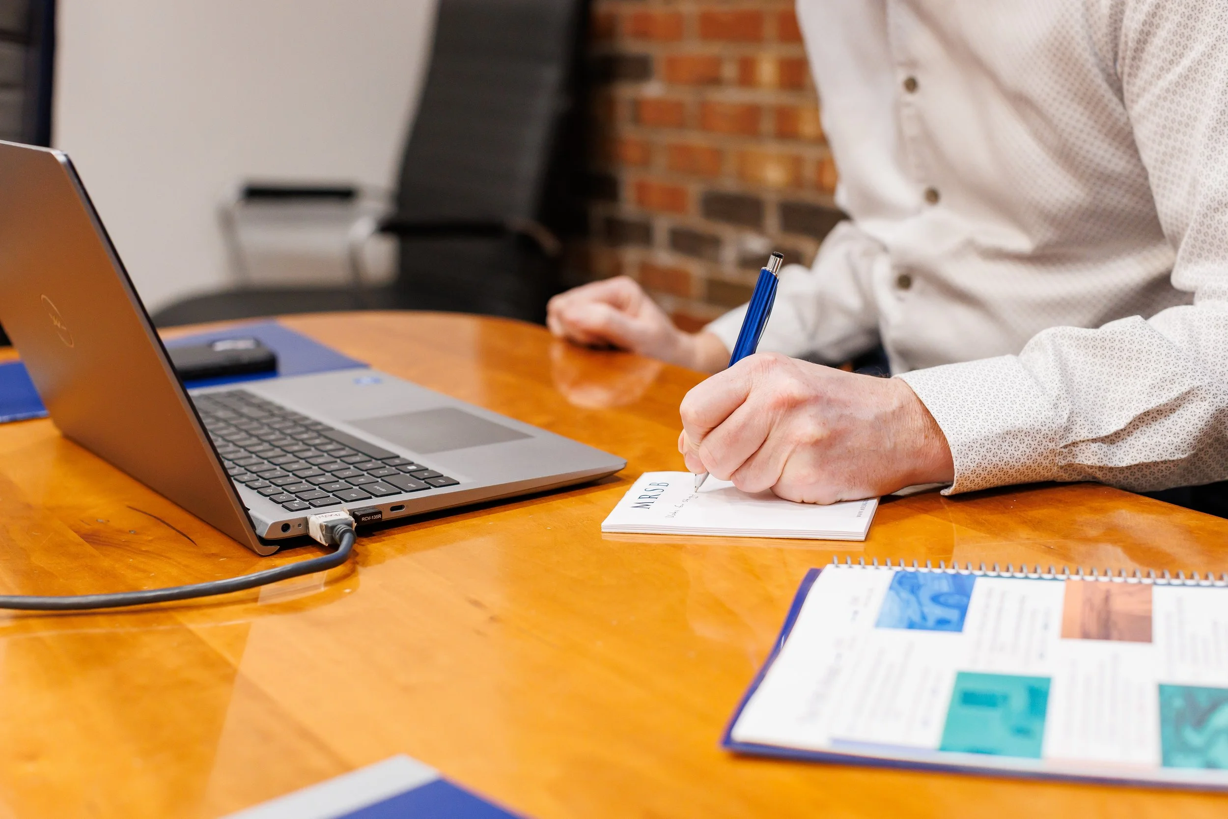 A person in a white shirt is sitting at a wooden table, writing on a notepad with a blue pen. There is a silver laptop in front of them, connected to a power cord, and some printed materials and notebooks on the table. The background has a brick wall and black chairs.
