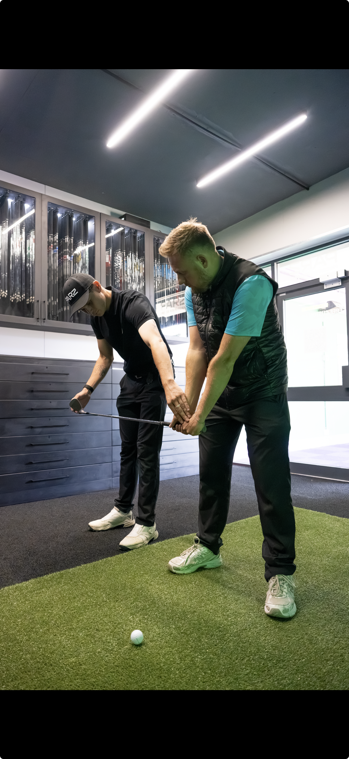 Two men practicing golf indoors; one is helping the other; both focus on a golf ball on a green mat.