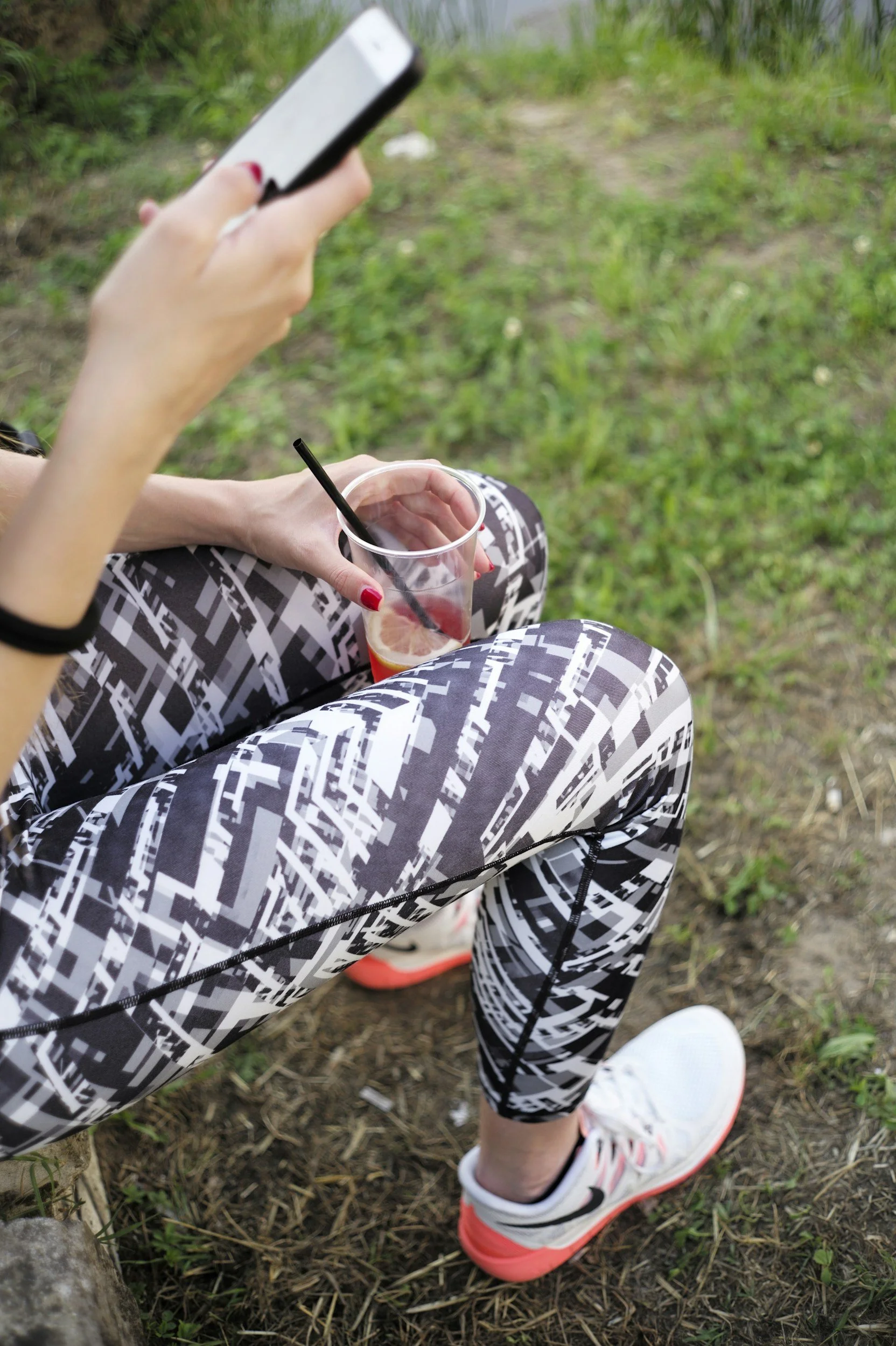 A woman sitting outdoors on grass in athletic leggings and sneakers, holding a smartphone in one hand and a clear cup of iced beverage with a straw in the other.