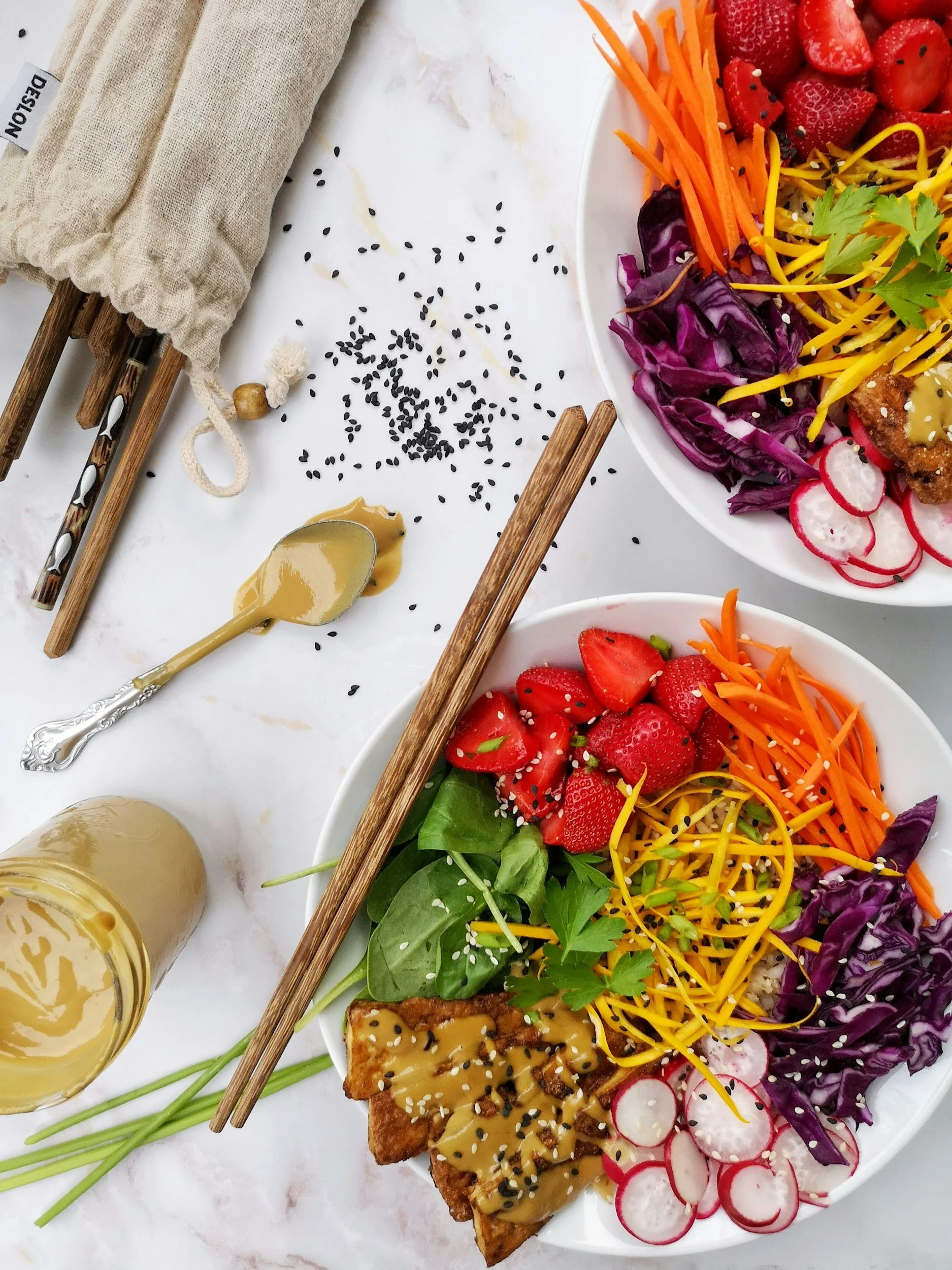 Veggie bowls with strawberries, shredded carrots, purple cabbage, radishes, greens, and tofu with sauce, chopsticks, and sesame seeds on white surface.