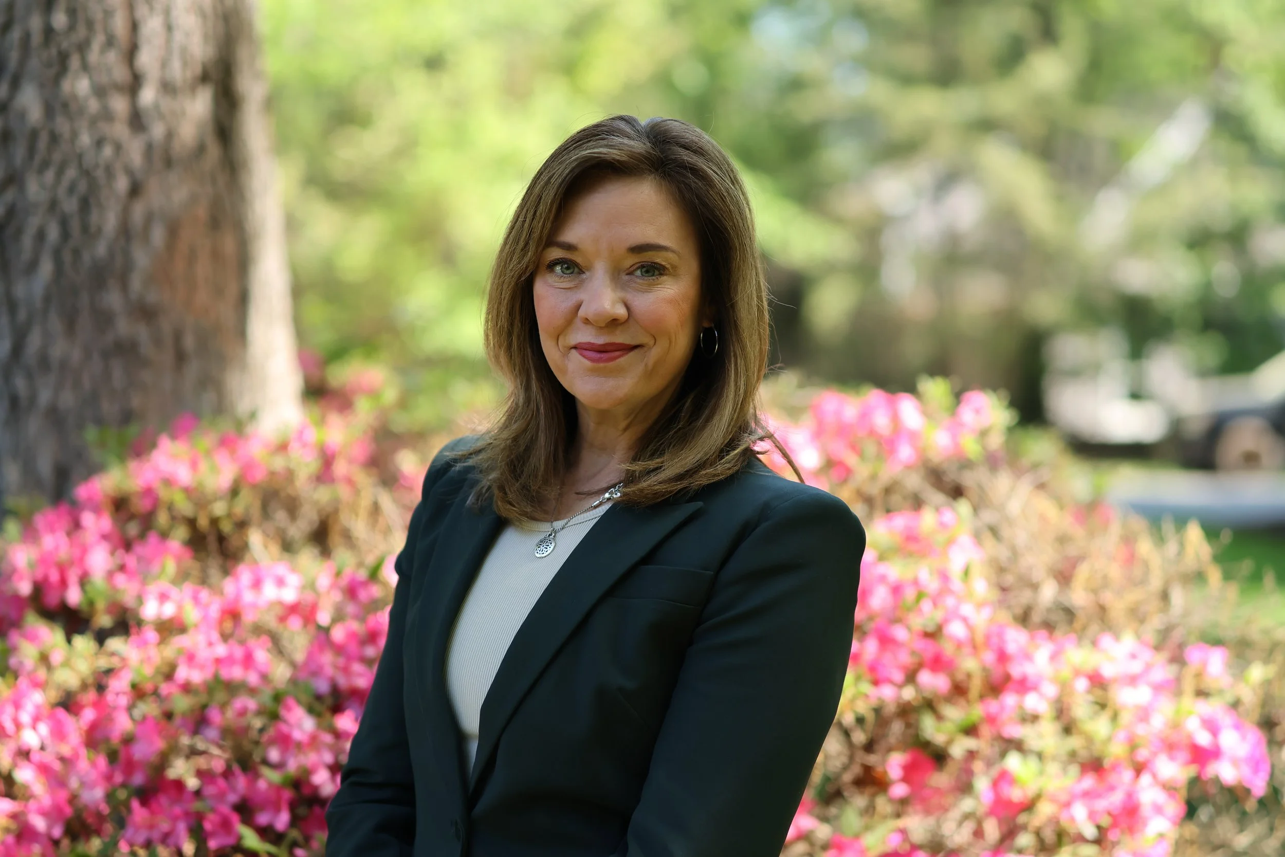 A woman with shoulder-length brown hair and a black blazer standing outdoors with pink flowers and green trees in the background.