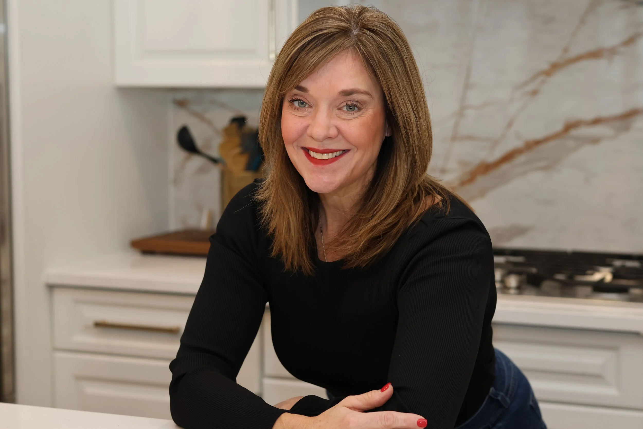 A smiling woman leaning on a kitchen counter with white cabinets and marble backsplash.