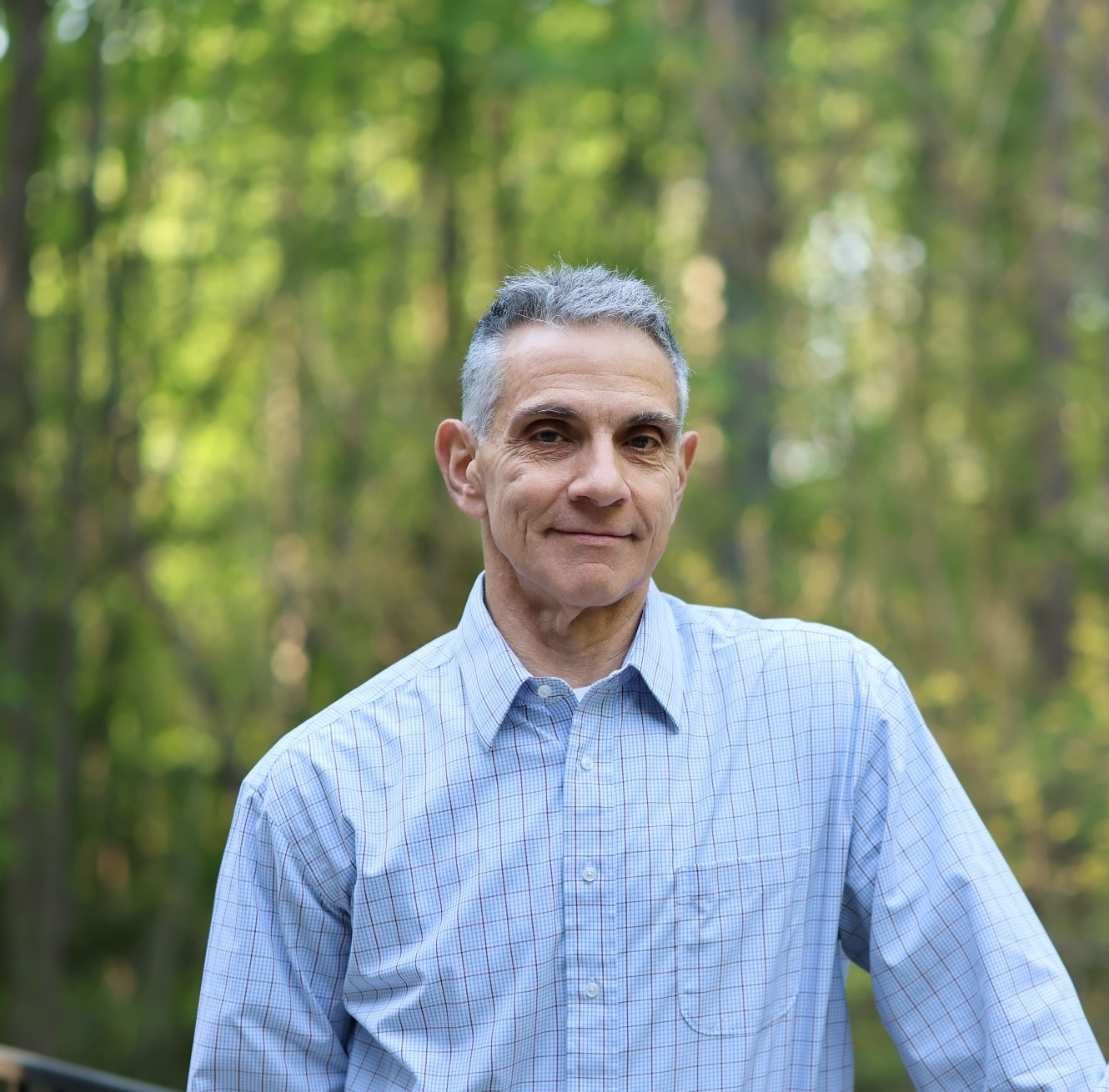 A middle-aged man with gray hair and a slight smile, wearing a light blue checkered shirt, standing outdoors in a forest with green trees in the background.