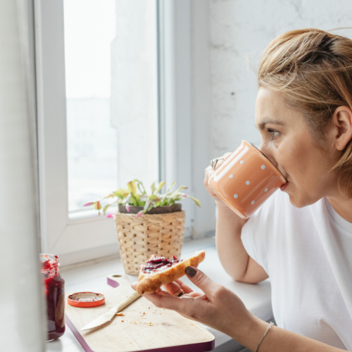 A woman with short blonde hair drinking from a pink polka dot mug while holding a slice of toast with jam.