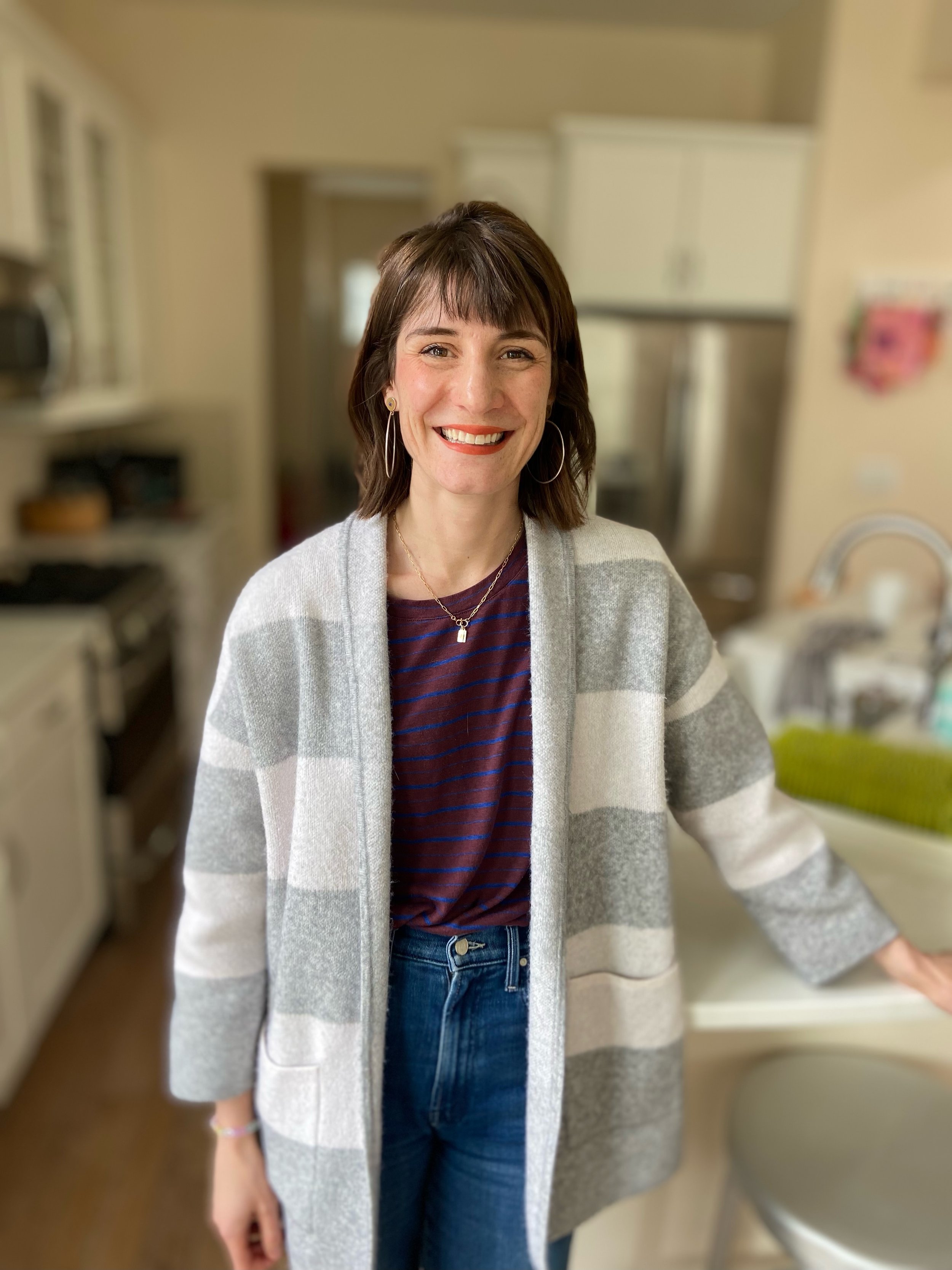 A woman standing in a kitchen, wearing a gray-striped cardigan, navy striped shirt, and jeans, smiling at the camera.