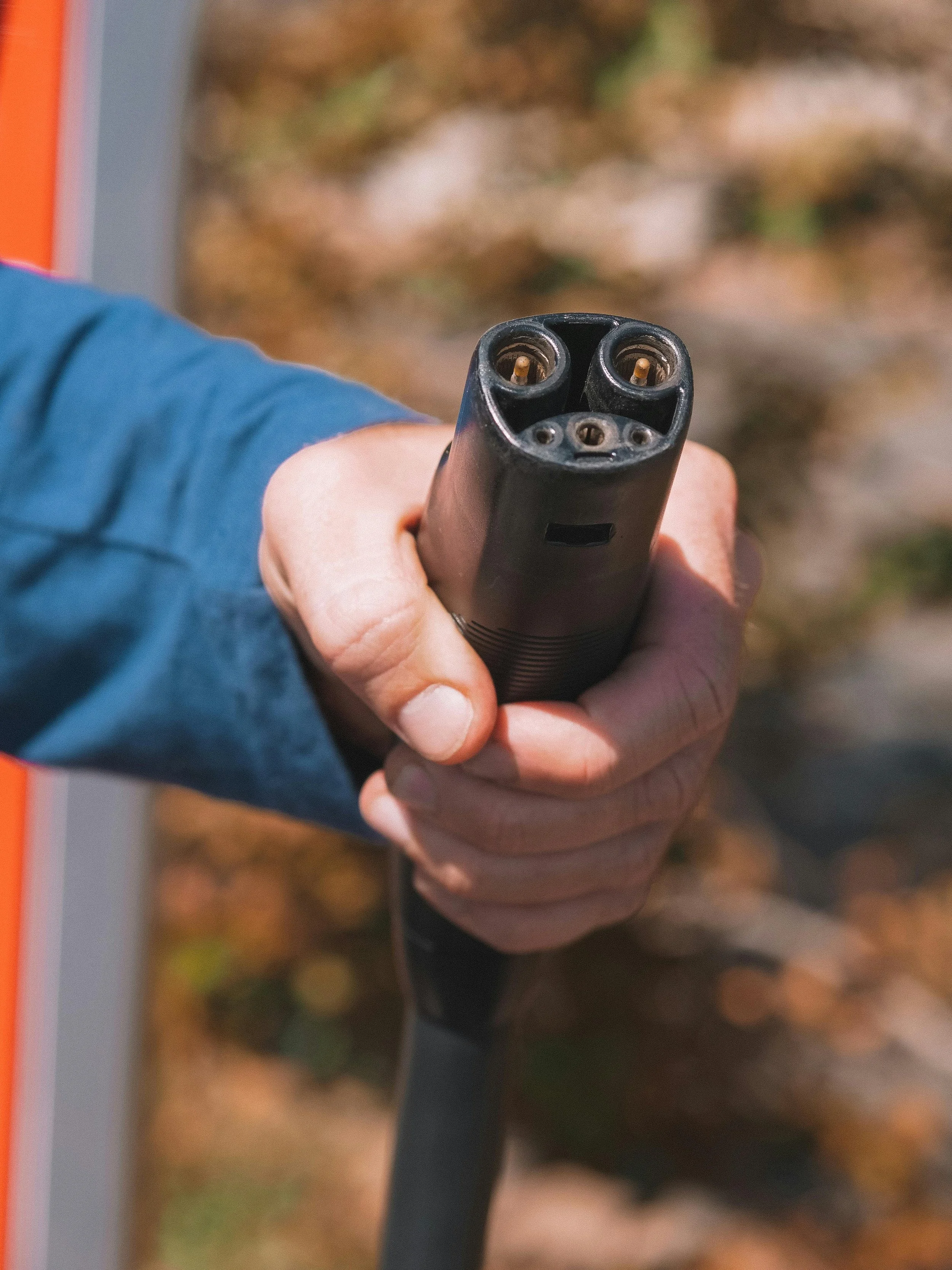 Close-up of a person's hand holding an electrical power connector, with trees and fall foliage in the background.