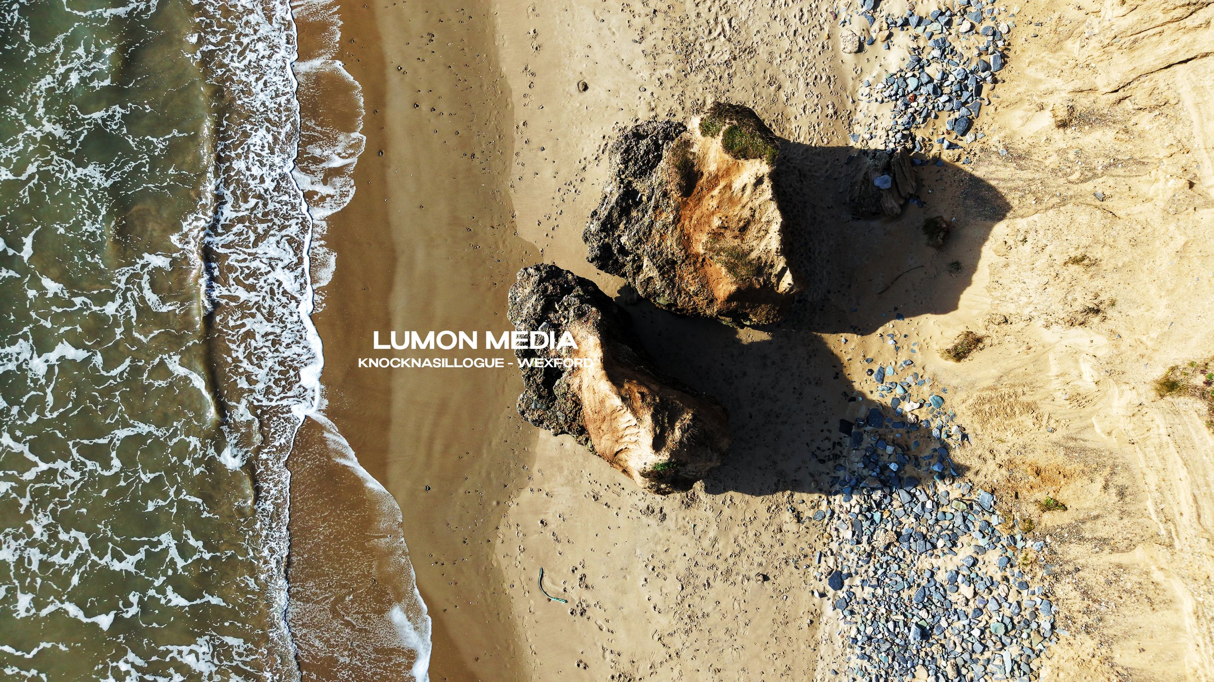 Bird's eye view of a sandy beach with two large rocks casting shadows, small rocks scattered nearby, and waves crashing onto the shoreline.