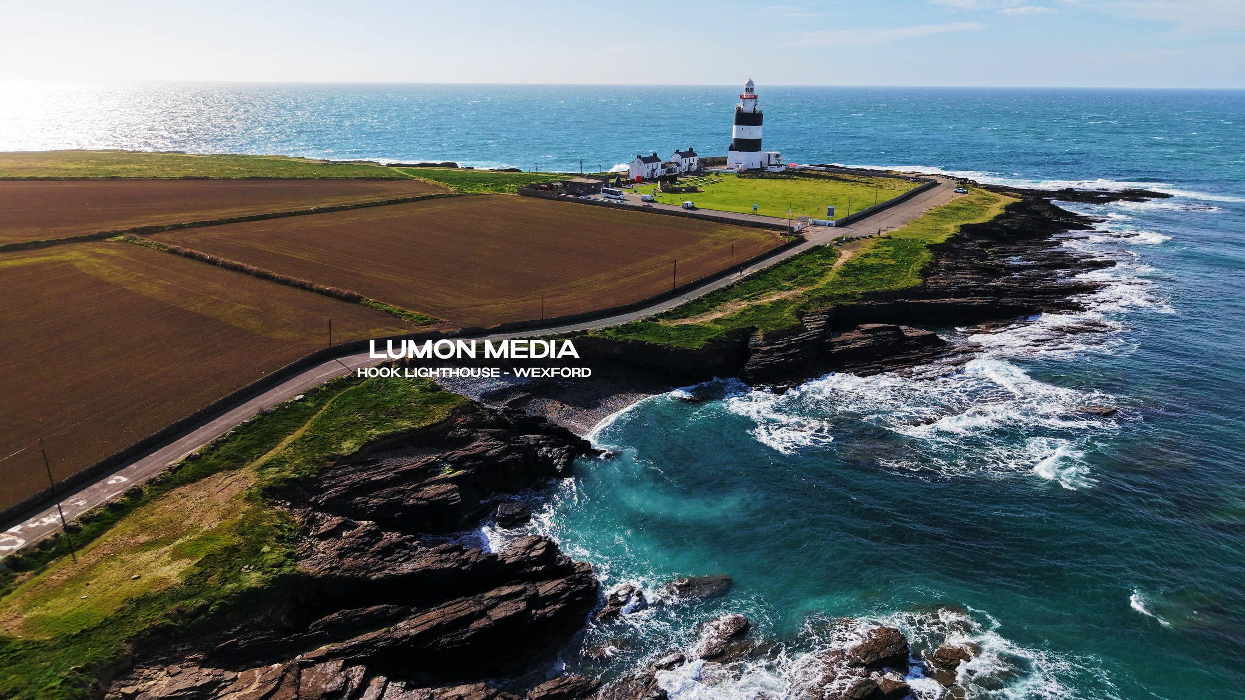 Aerial view of Hook Lighthouse on the coast of Wexford, Ireland, with the lighthouse, coastal cliffs, waves crashing against rocks, and farmland nearby under clear skies.