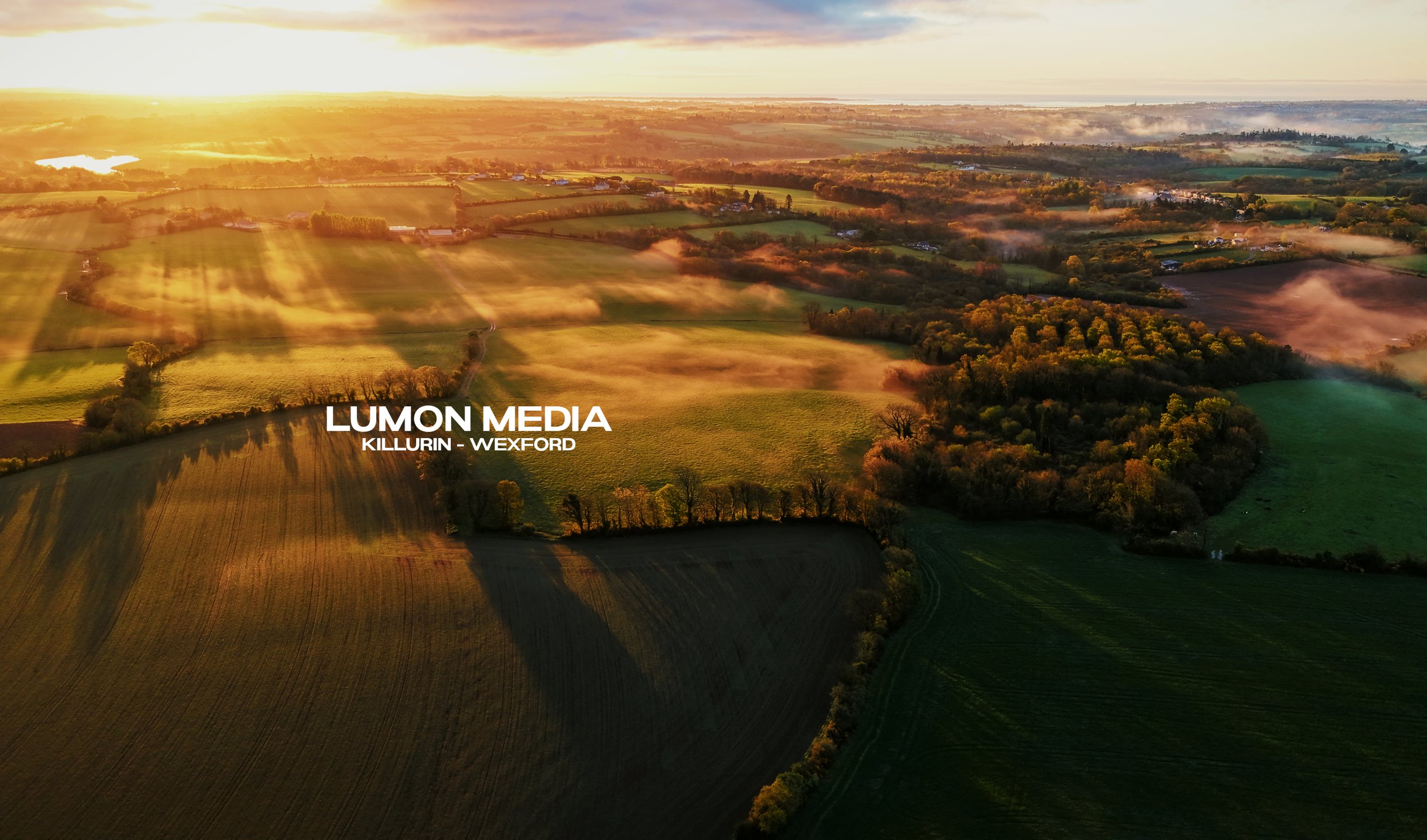 Aerial view of a rural landscape with fields, trees, and mist at sunrise, with shadows cast by the trees and the sky showing a mix of clouds and sunlight. Text overlay reads 'LUMON MEDIA KILLURIN - WEXFORD'.