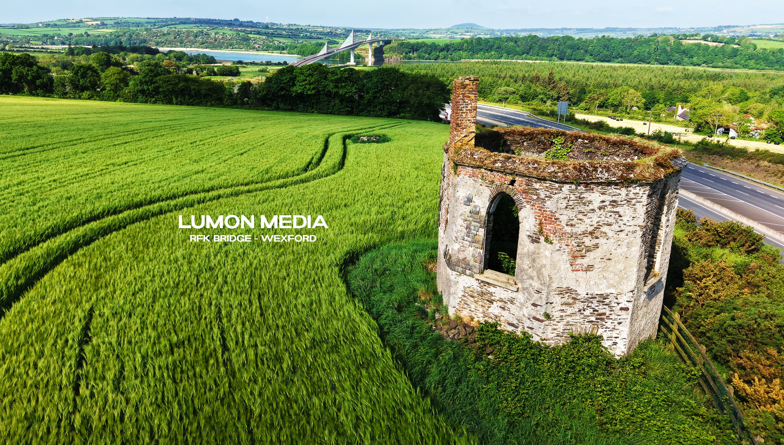 Green fields with tall grass and winding paths, old stone tower, highway with traffic, bridge in the distance under a blue sky.