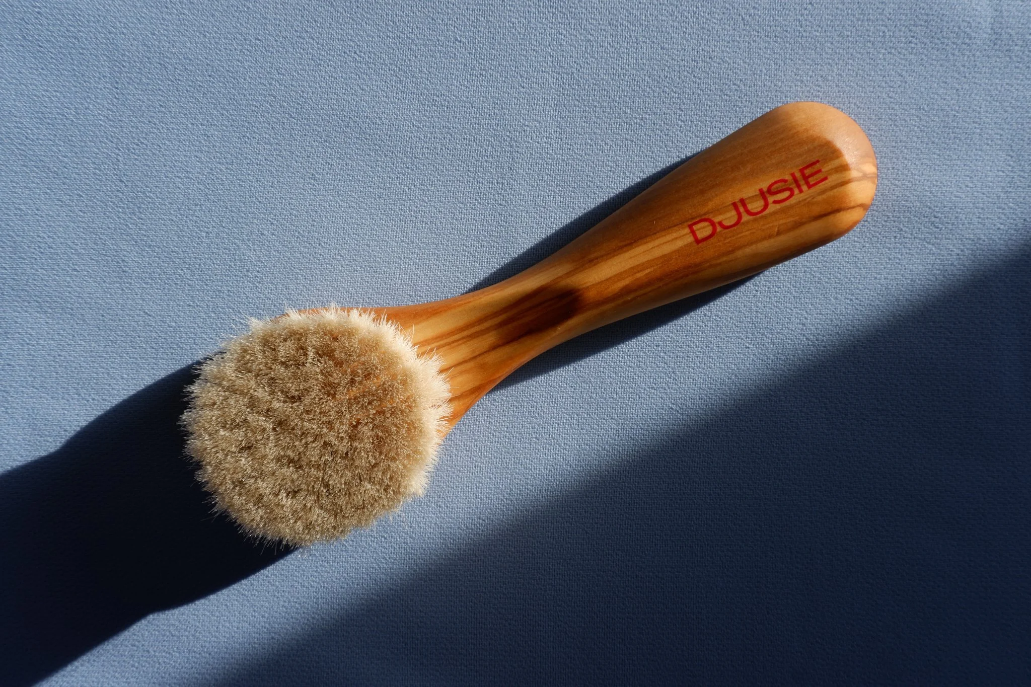 A wooden brush with a round, beige bristle head placed on a blue surface, with a shadow cast to the right. The handle has red text that reads 'DJUSIE'.