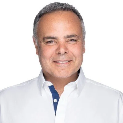 A man with short gray hair smiling, wearing a white shirt with a contrasting blue button placket, against a plain white background.
