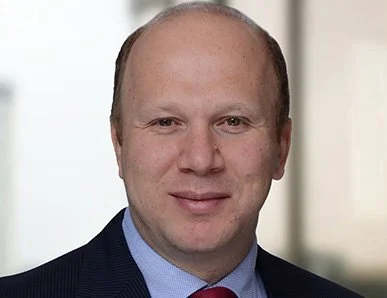A professional headshot of a man in a suit and tie, smiling, with a blurred office background.