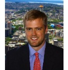 A man with short brown hair and a beard, wearing a dark suit, blue shirt, and red tie, standing outdoors with a city skyline and blue sky in the background.