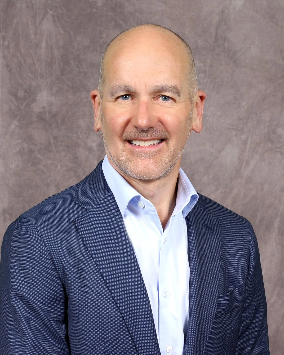 Headshot of a middle-aged man with a smile, wearing a dark blue suit and a light blue dress shirt, against a neutral background.