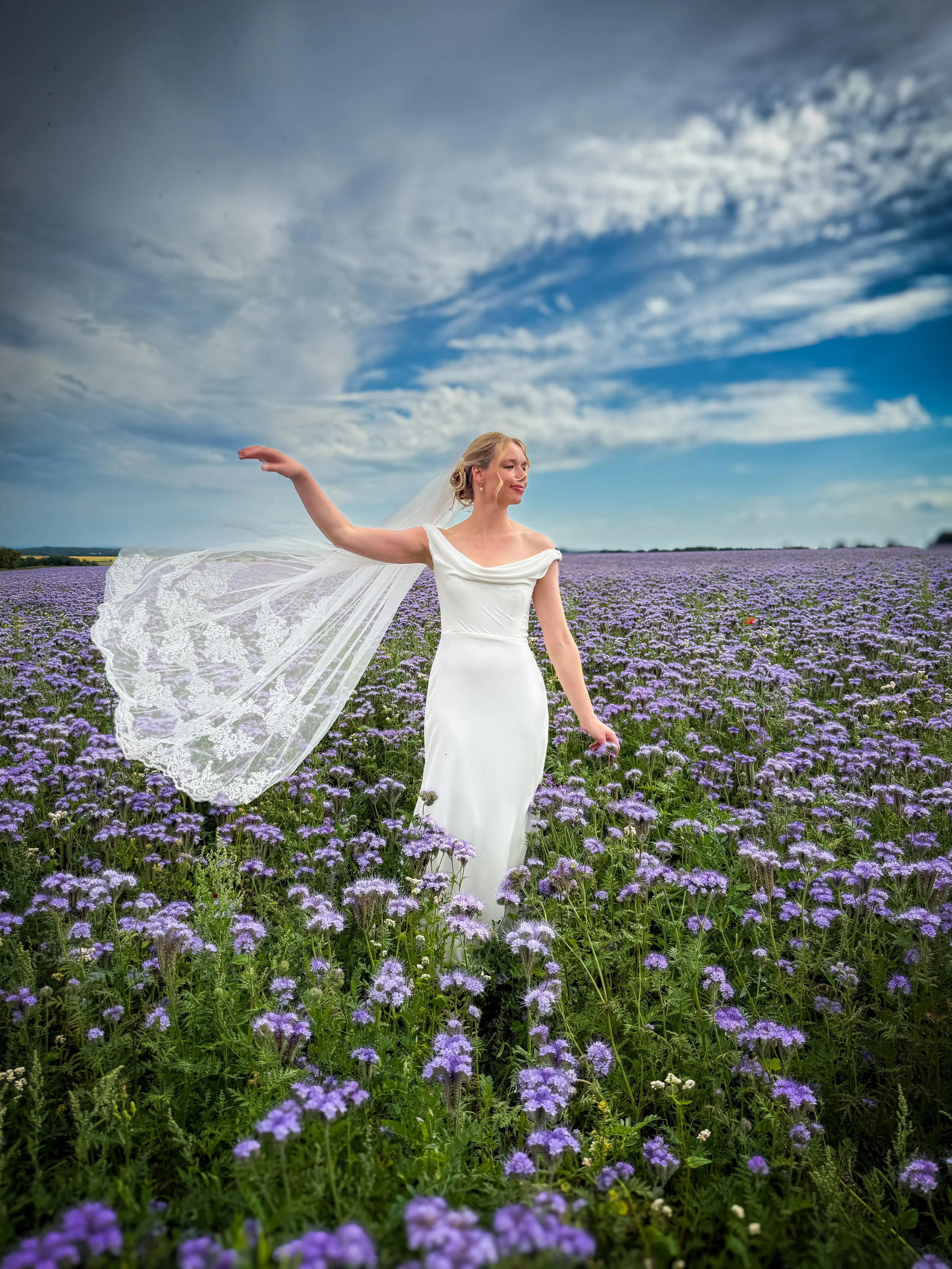A woman in a white dress standing in a field of purple flowers under a partly cloudy sky, with one arm extended outward and a gentle smile.