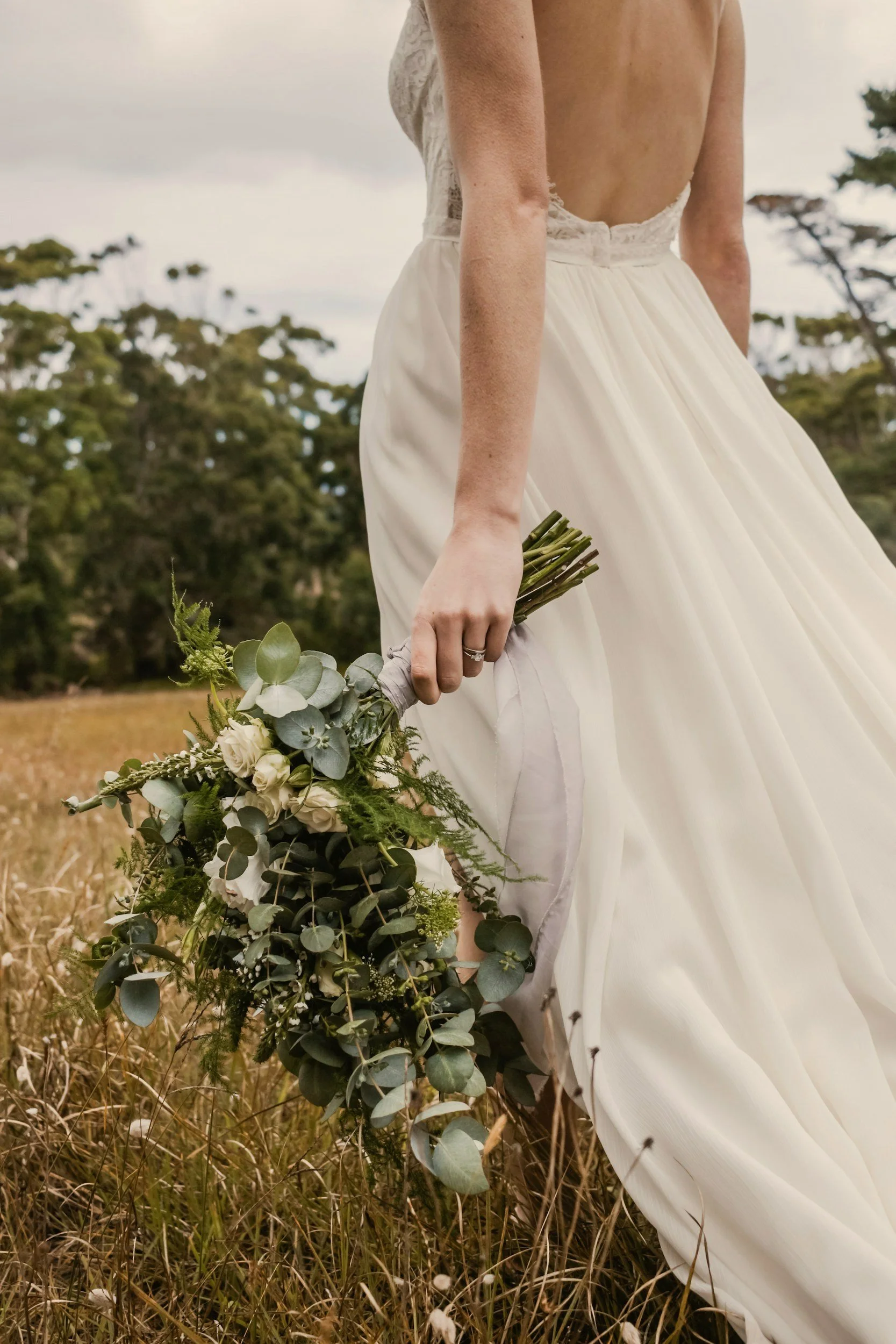 A bride in a white wedding gown holding a bouquet of greenery and white flowers in a field with trees in the background.