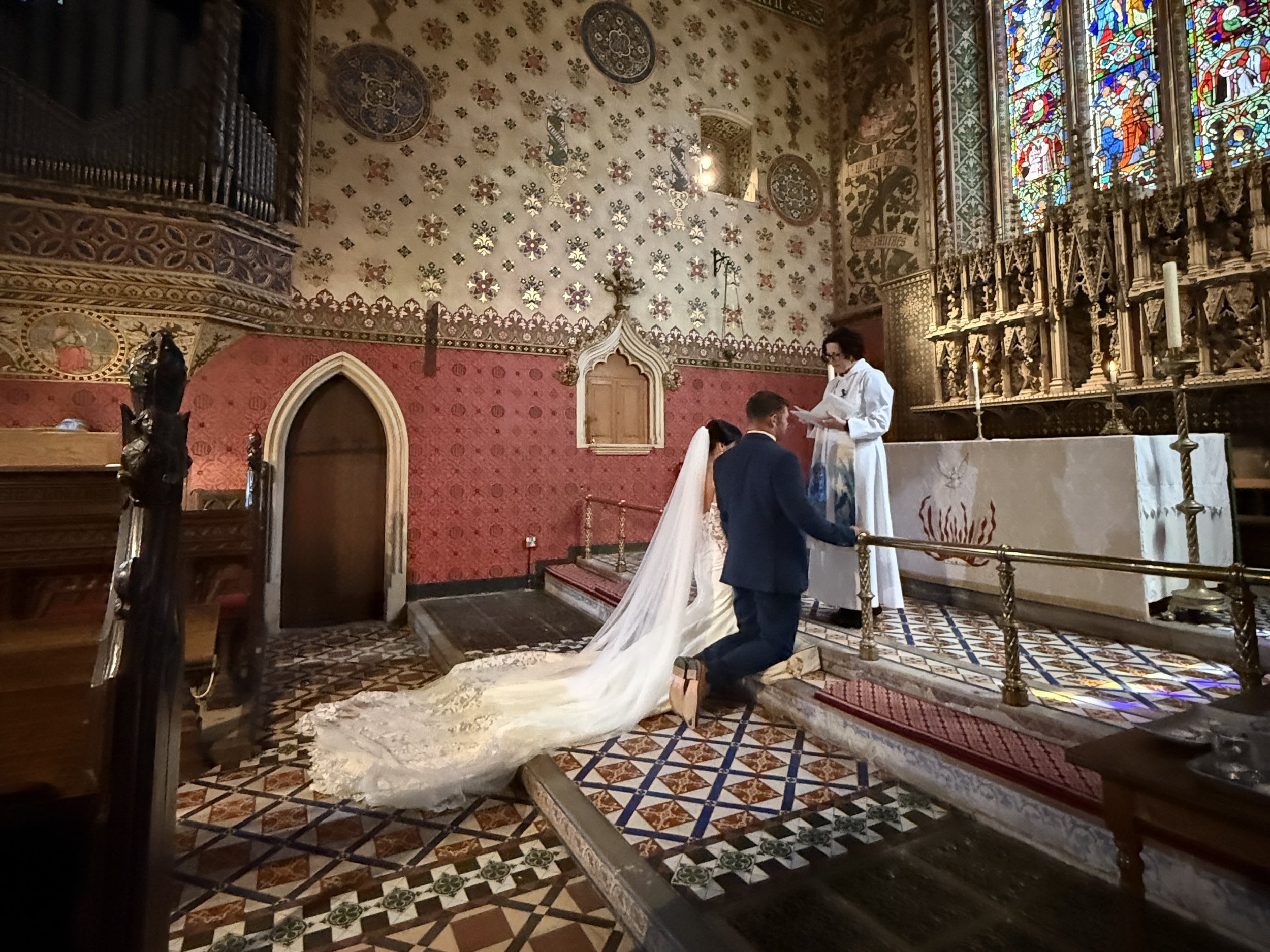 A wedding ceremony taking place inside a richly decorated church with stained glass windows and ornate woodwork. A bride and groom kneel before a clergy member as the bride's long train flows down the steps.