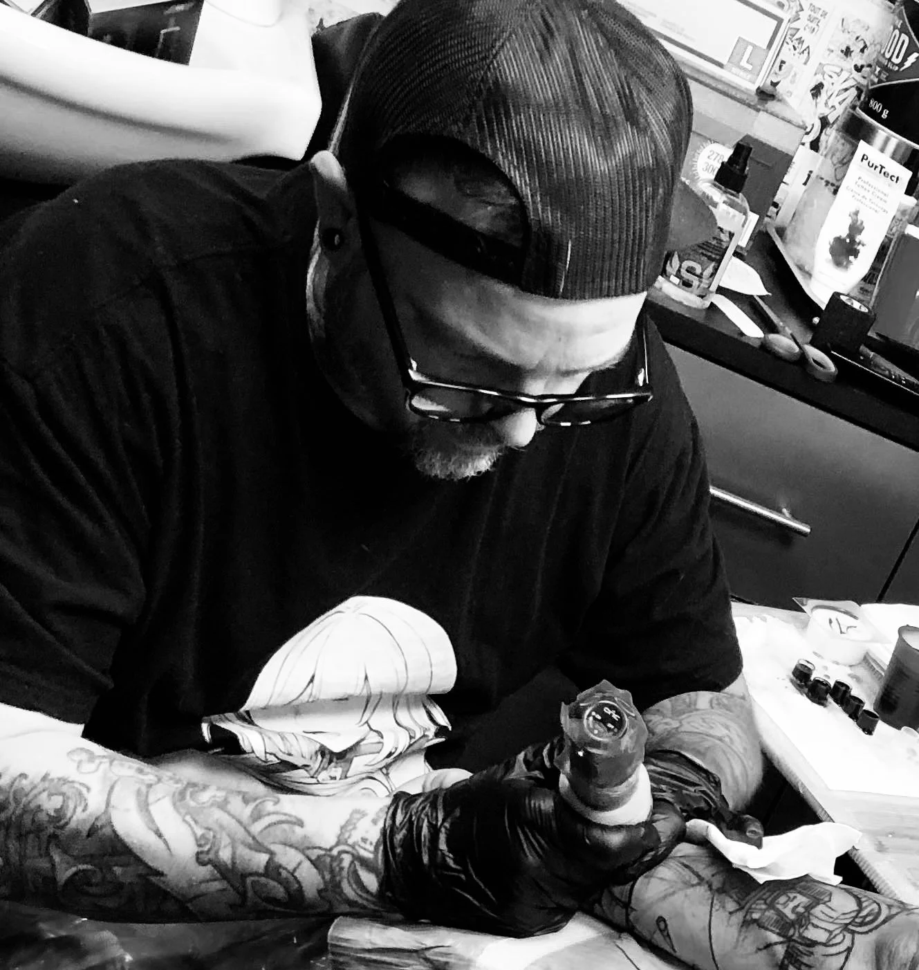 Un homme portant une casquette derrière et des lunettes, tatoué, qui se fait tatouer par un artiste dans un salon de tatouage, en noir et blanc.