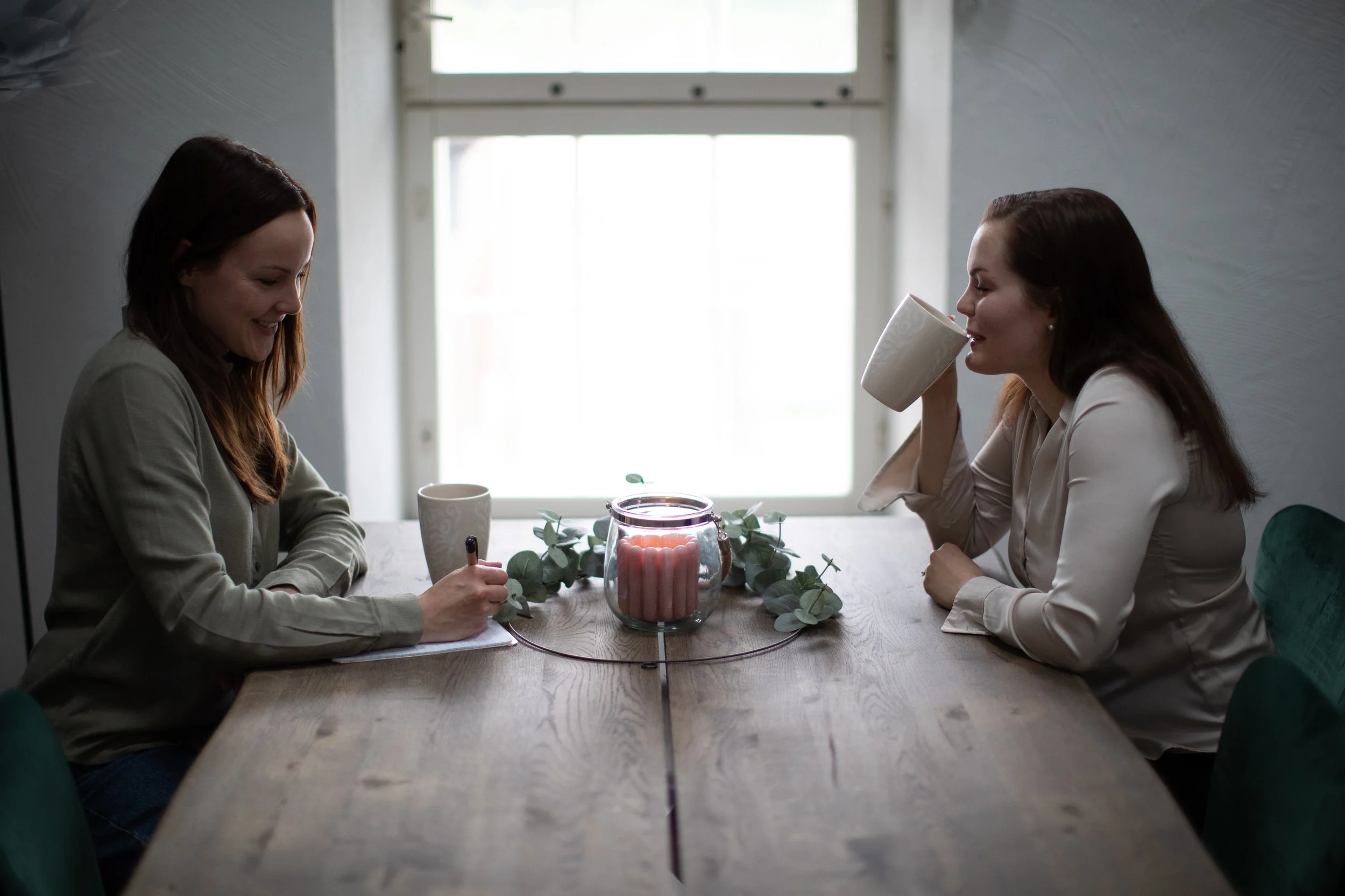 Two women sitting at a wooden dining table, one writing and the other drinking from a mug, with a candle and greenery in the center, in a room with a window in the background.