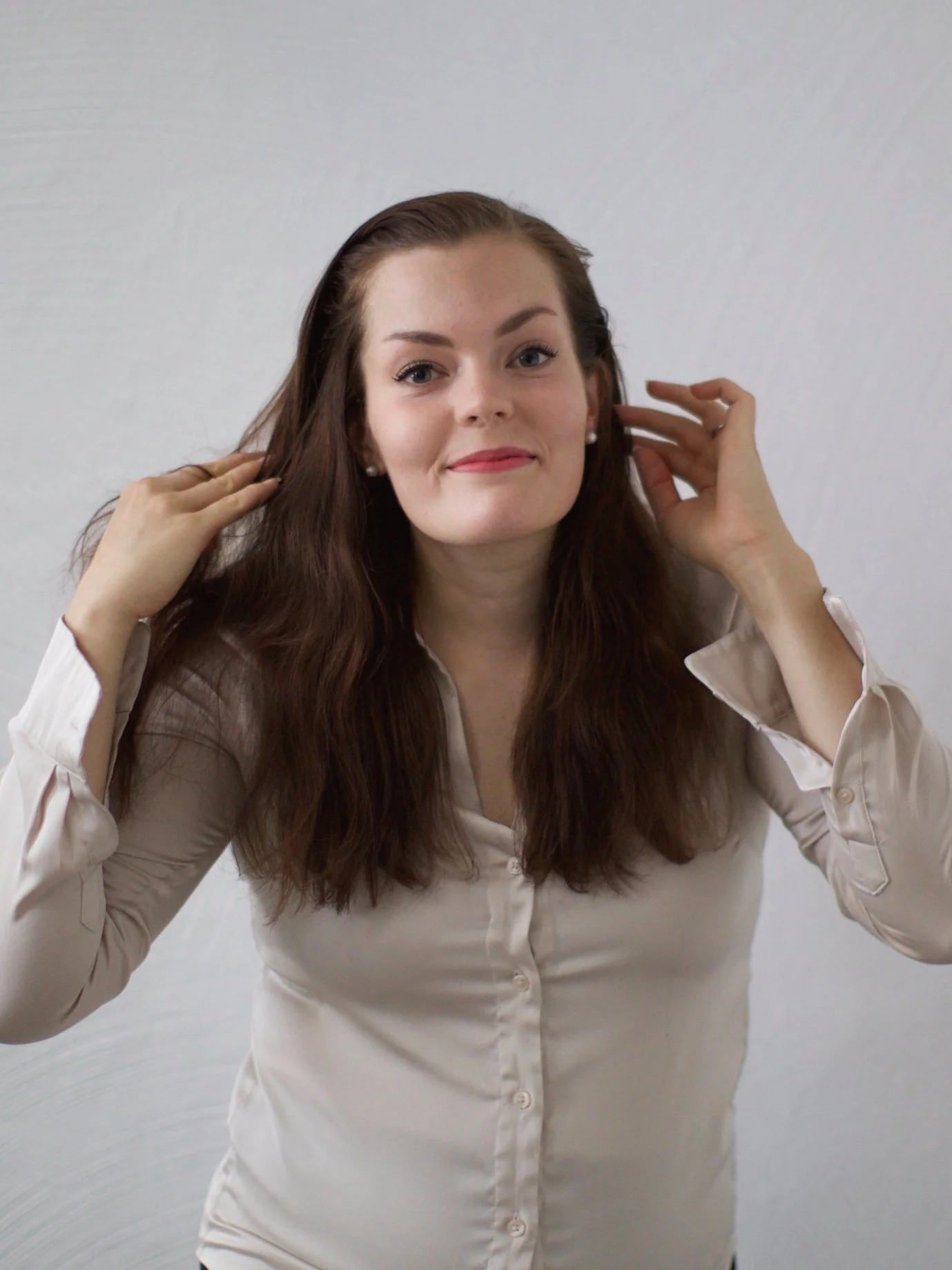 A woman with long brown hair, wearing a cream-colored button-up shirt, smiling and touching her hair.