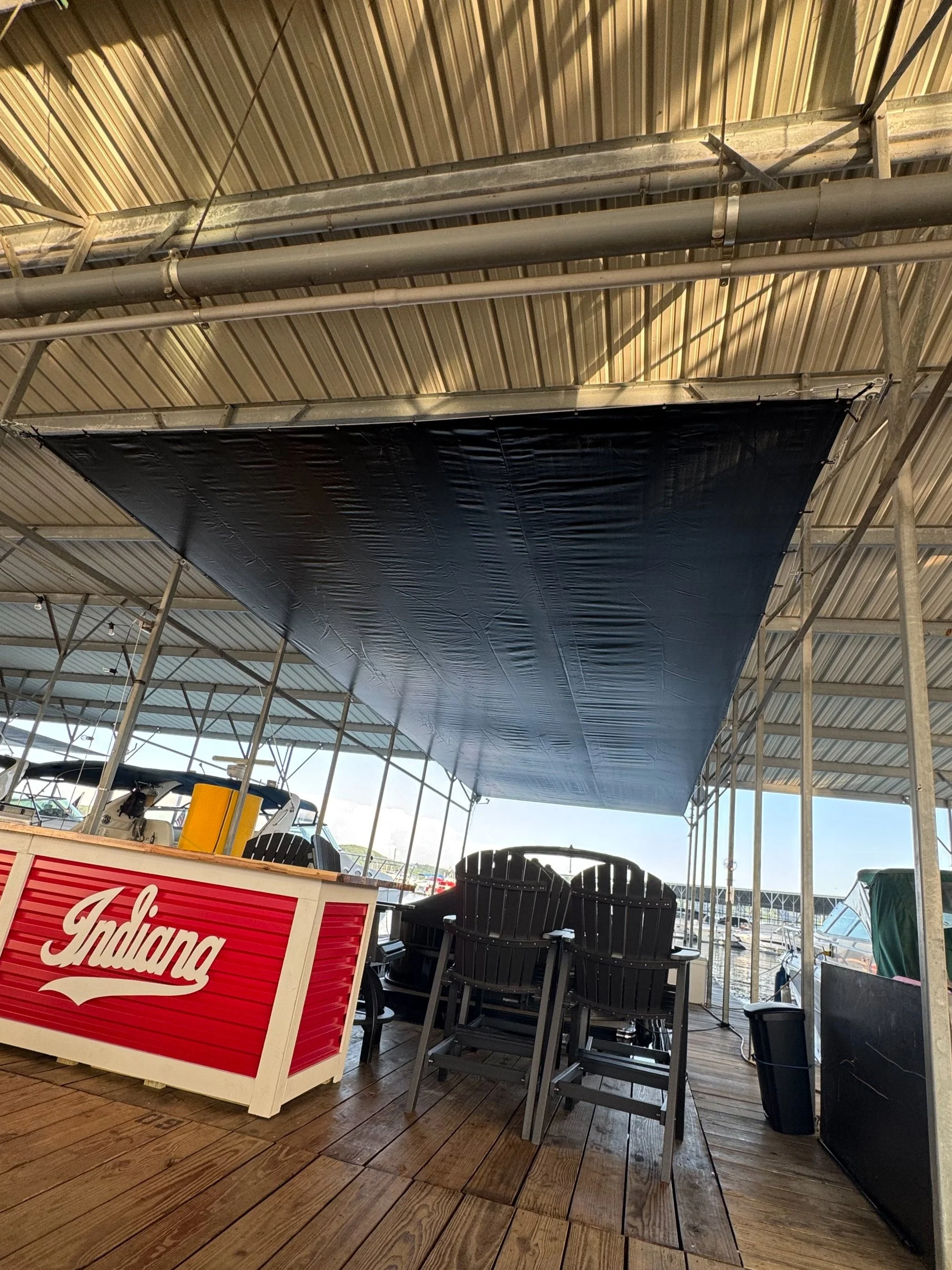 Boat dock with black shade sail, stacked black Adirondack chairs, red Indiana Bar sign, and boats in the background at a marina.