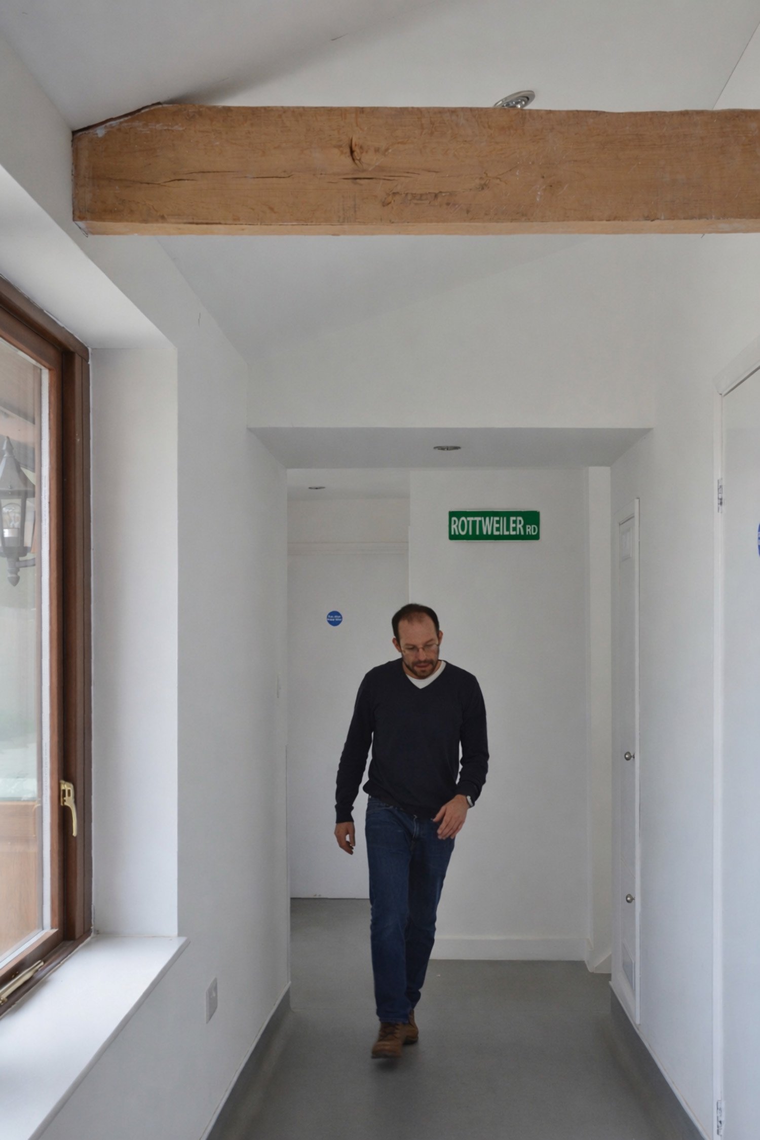 A man walking in a white corridor with a window on the left and a green street sign reading 'ROTTWEILER RD' in the background.