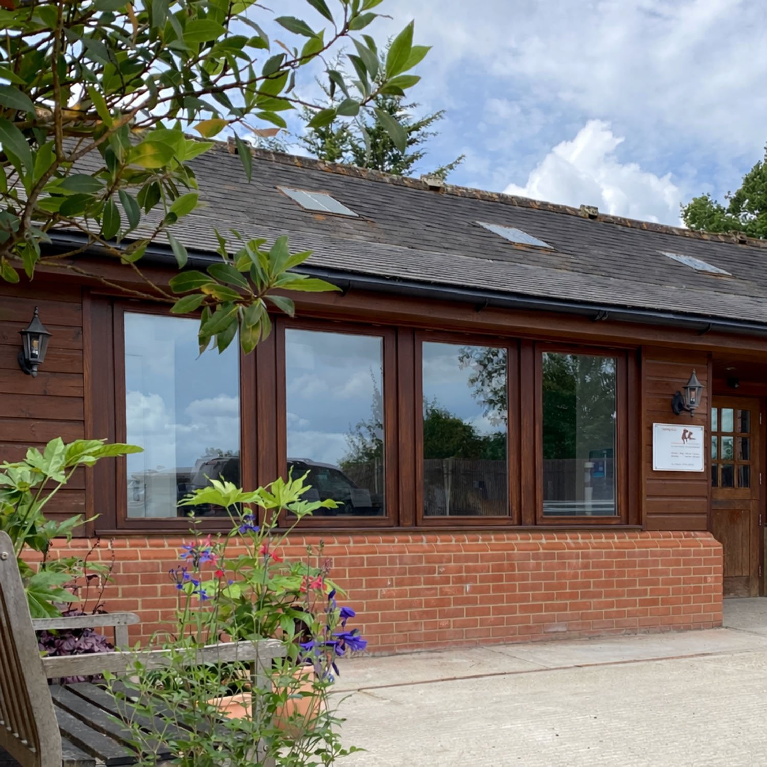 Exterior of a building with large windows, brick and wooden siding, surrounded by plants and flowers, with a cloudy sky overhead.