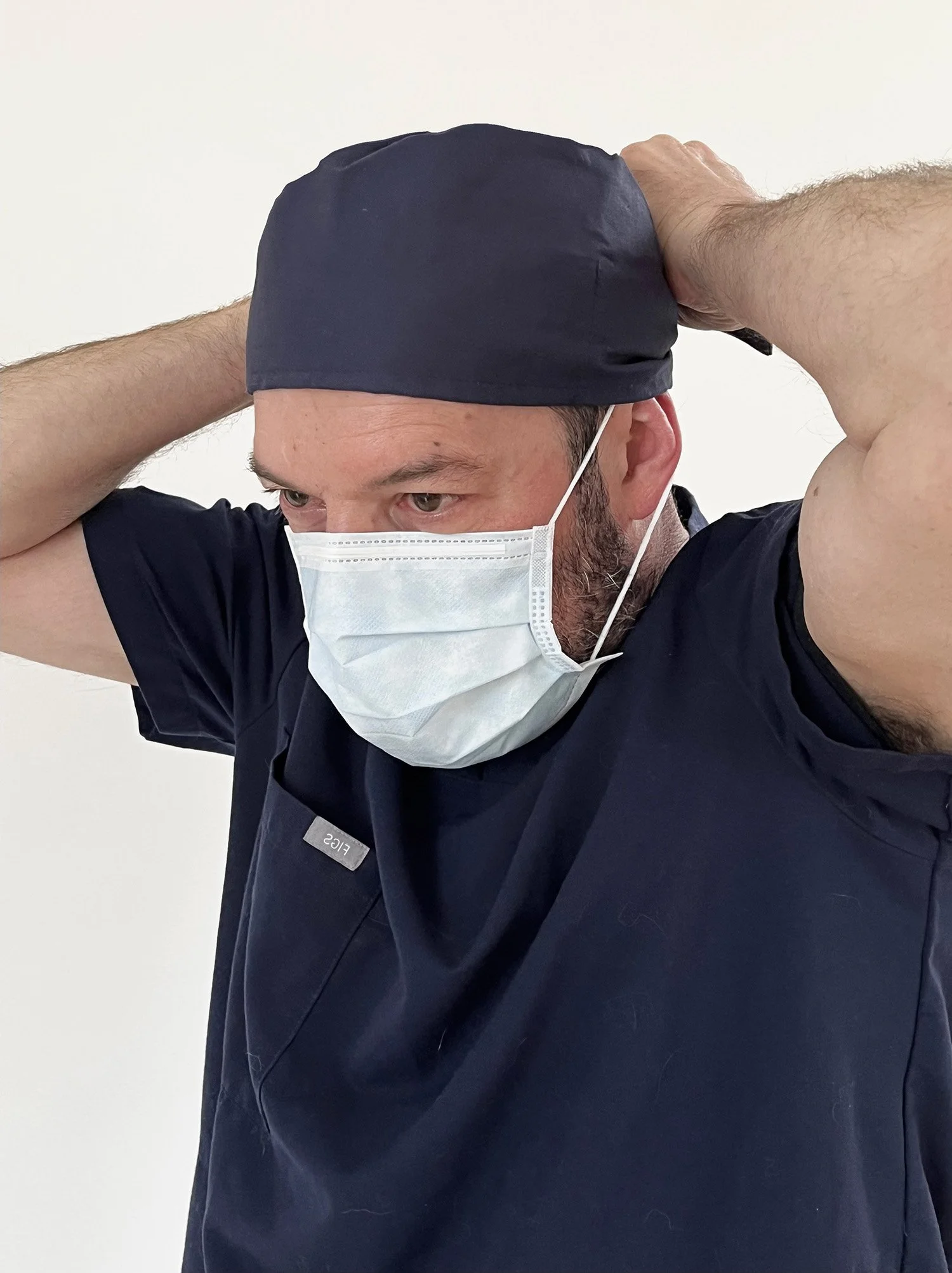 A healthcare professional in navy scrubs, a face mask, and a cap, adjusting the cap with his hands, against a plain white background.
