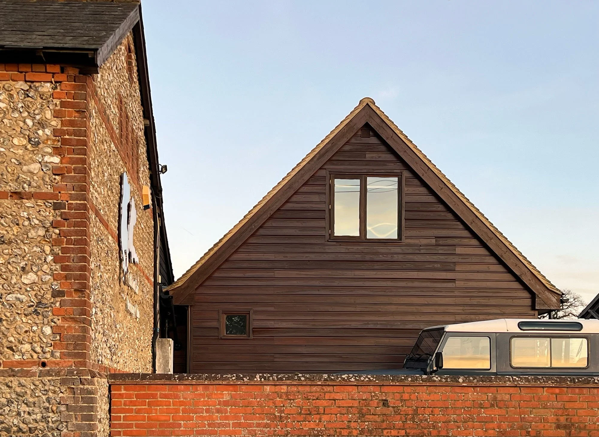 Buildings with a brick wall and a wooden house with a triangular roof, with a white vehicle parked in front.