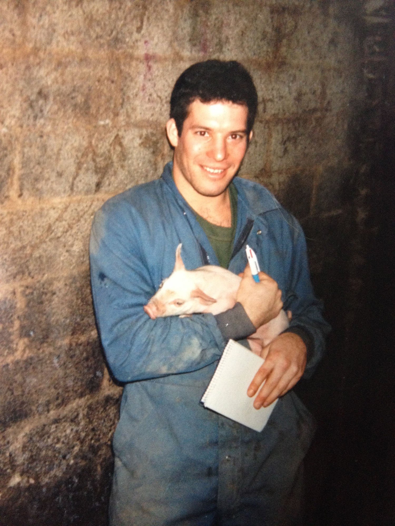 A young man with dark hair and a blue jacket holding a piglet with its eyes closed, standing against a stone wall.