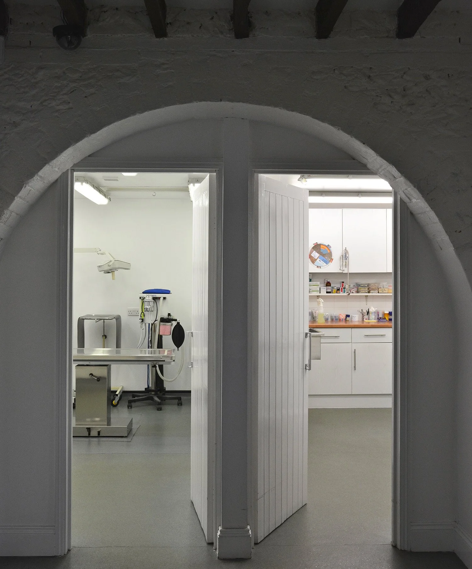 View into a medical or veterinary clinic, showing a treatment room with an examination table and medical equipment on the left, and a storage and supply area with cabinets and various supplies on the right.