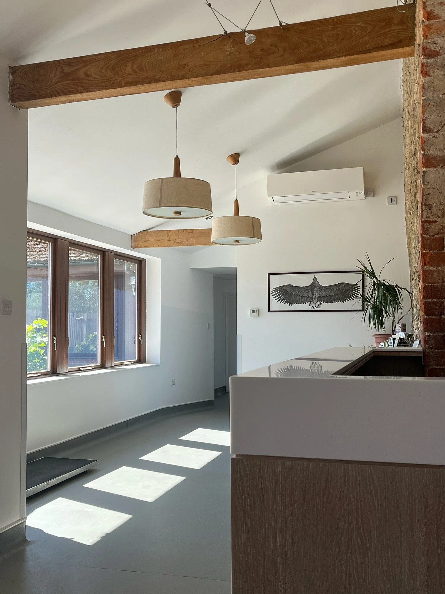 Sunlight coming through wooden-framed windows shining onto a light-colored floor in a modern, minimally decorated living space with white walls, ceiling-mounted air conditioning unit, hanging beige lamps, a framed bird illustration, potted plant, and a brick wall detail.