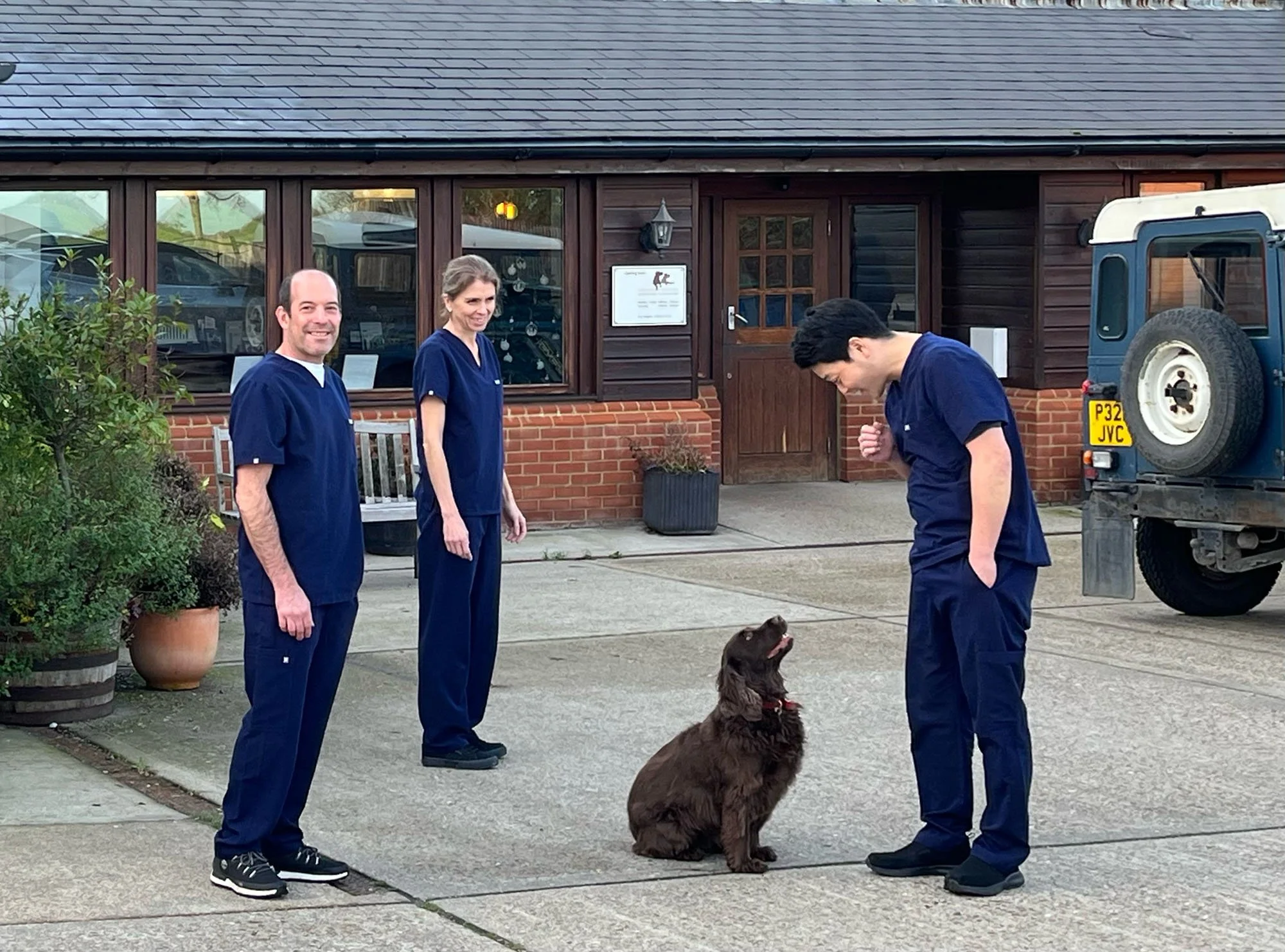 Four veterinary professionals, three men and one woman, in navy blue scrubs, outside a building. One brown dog sits on the ground, looking up at a young man with black hair, who is a few feet away. The man gestures with his right hand and looks down at the dog. The other two professionals stand nearby, smiling, observing the scene. A white vehicle and a brick building with windows and a door are in the background.