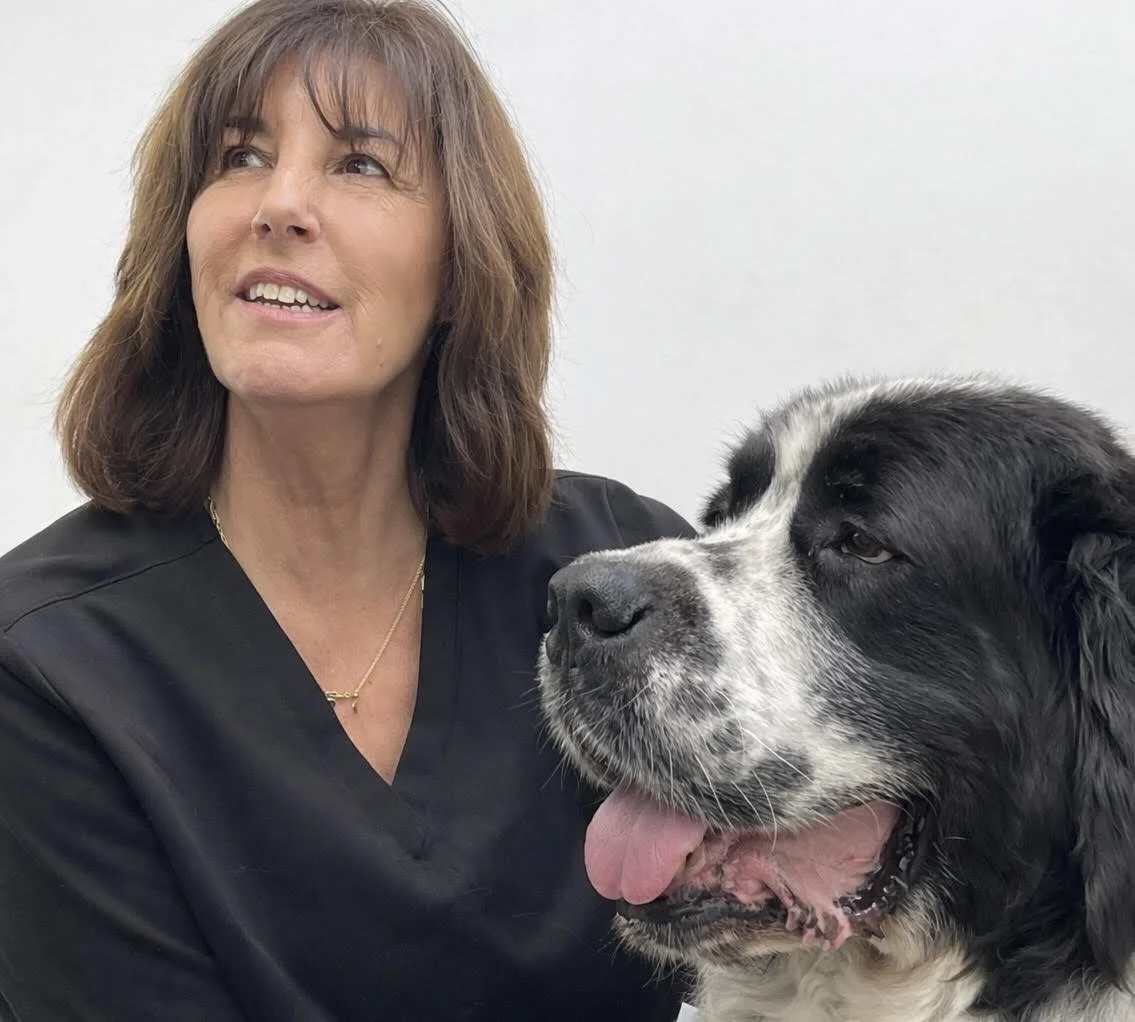A woman with brown hair smiling and looking up, wearing a black jacket and necklace, with a black and white dog resting its head on her shoulder.