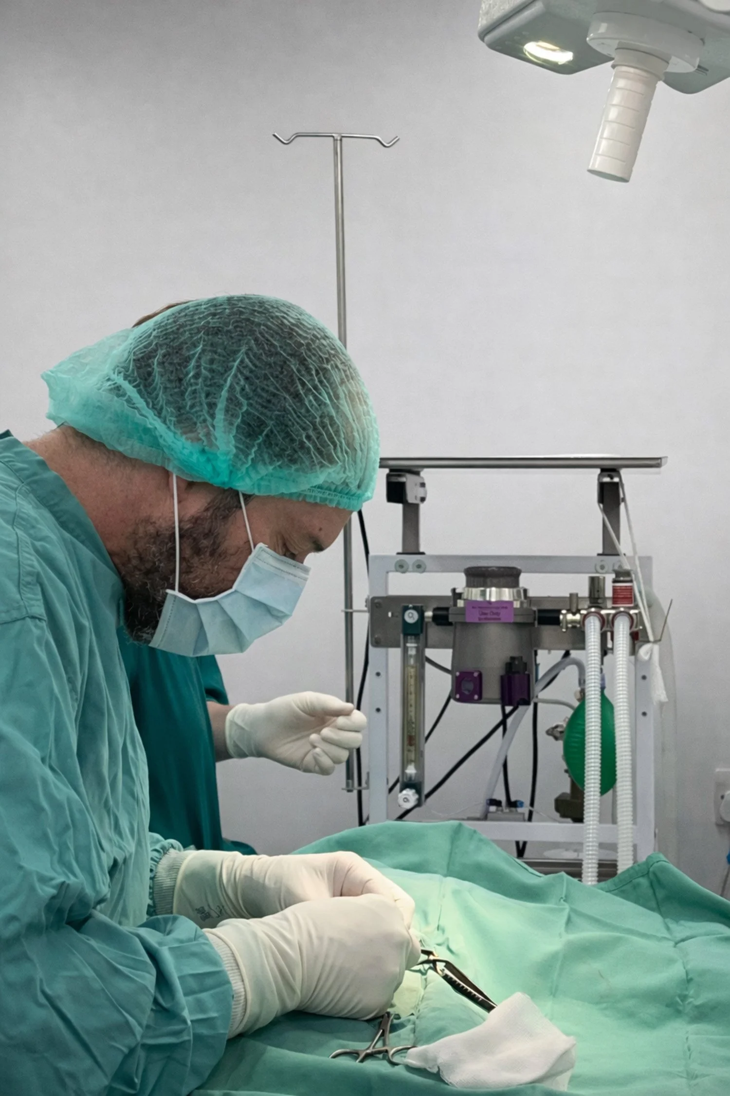 A surgeon dressed in scrubs, mask, cap, and gloves performing surgery in an operating room with medical equipment around.