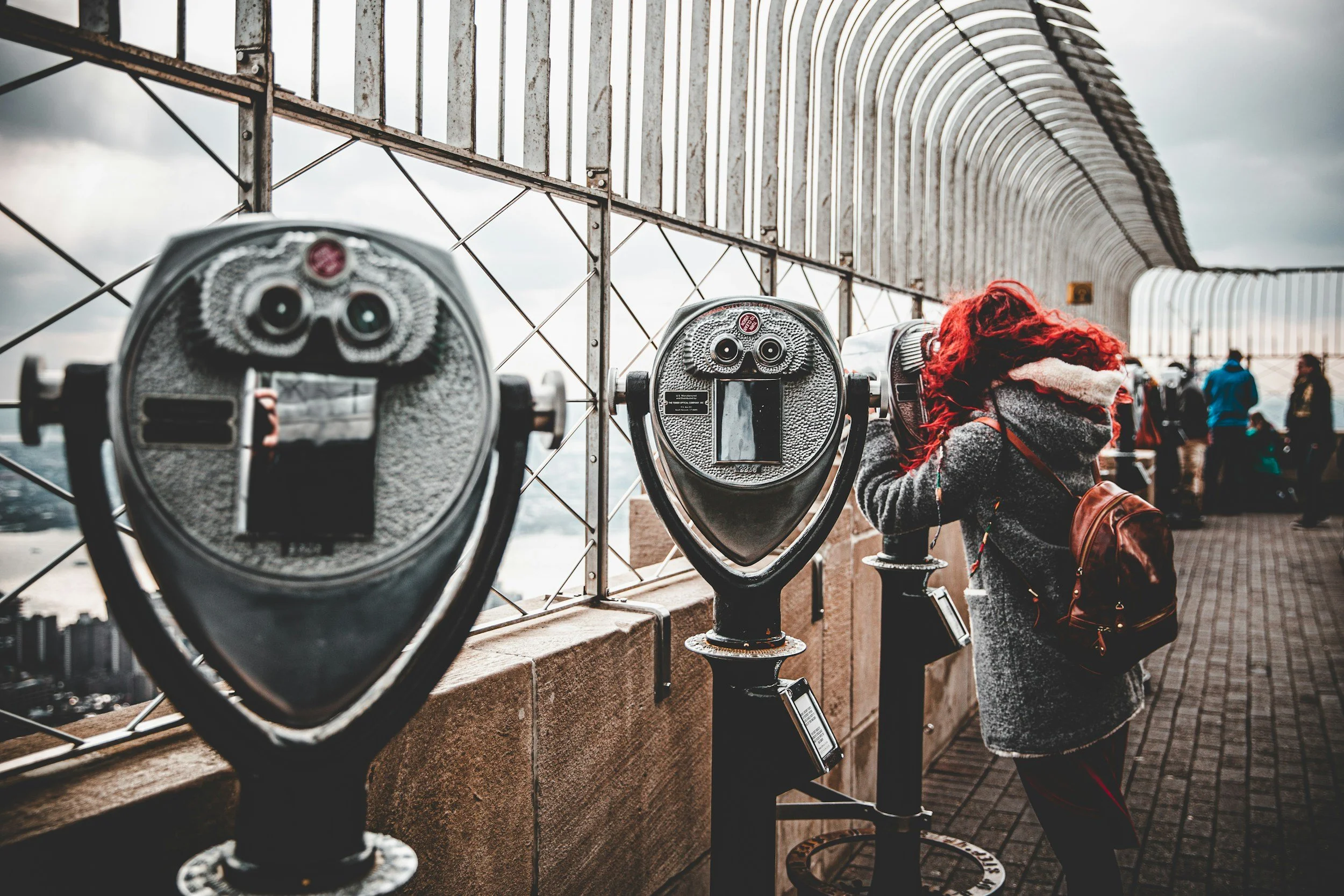 View of coin-operated binoculars on a viewing platform with a person wearing a gray coat and red hair looking through one of the binoculars, overlooking a cityscape on a cloudy day.