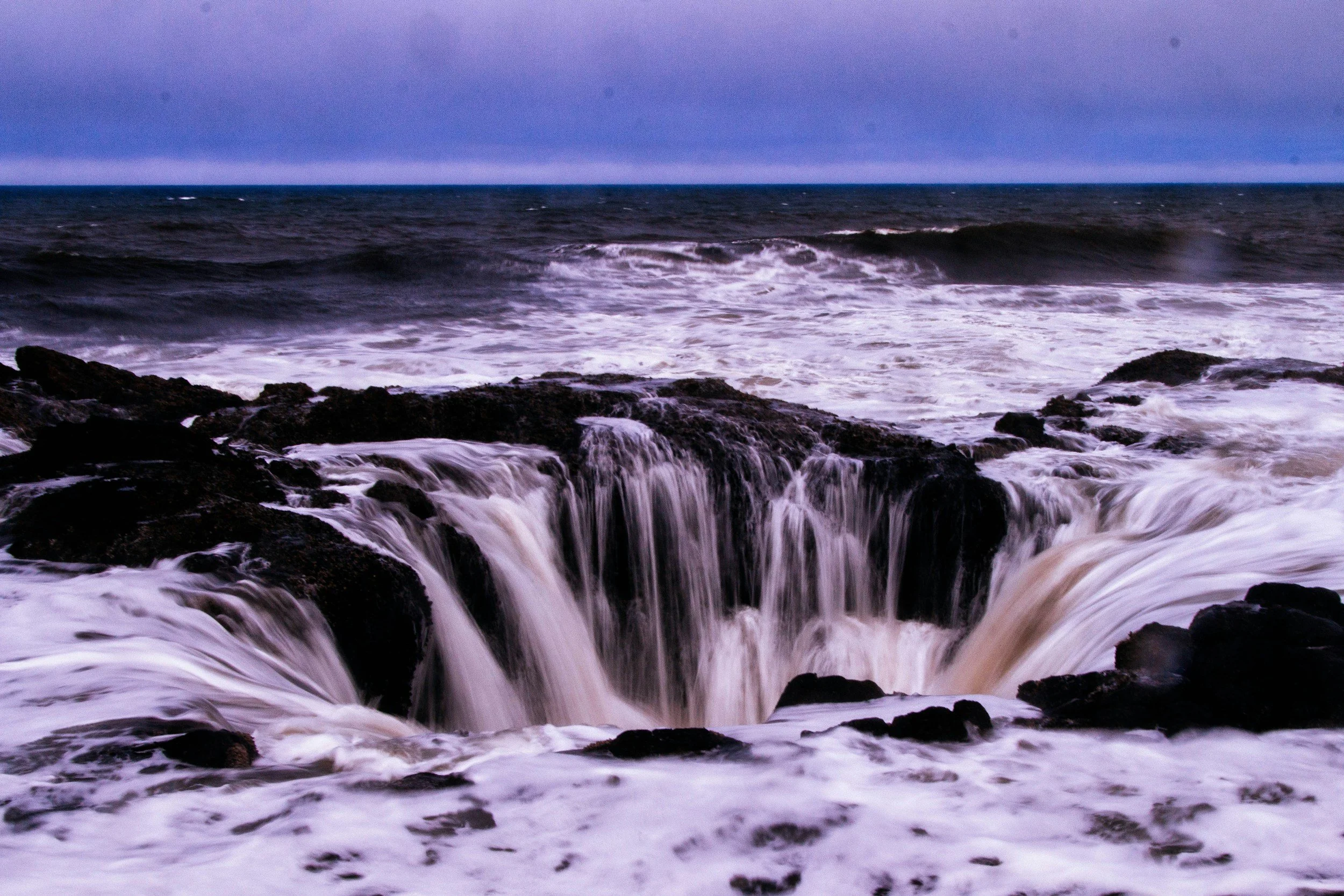Ocean waves crashing over rocks during sunset or sunrise with dark clouds overhead.