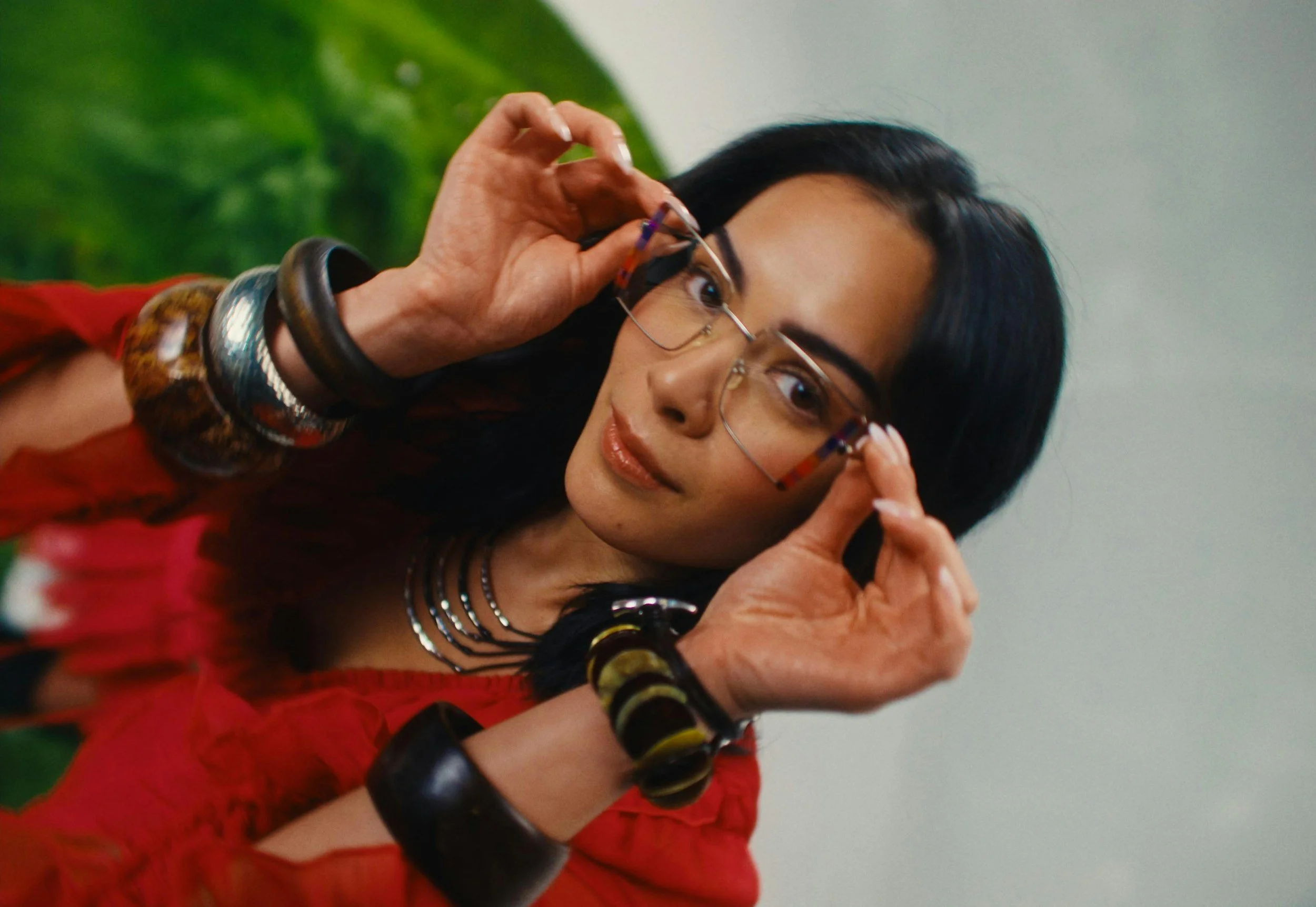 Young woman with black hair and tan skin putting on glasses, wearing a red shirt and layered necklaces, with multiple bracelets on both wrists, standing against a green background.