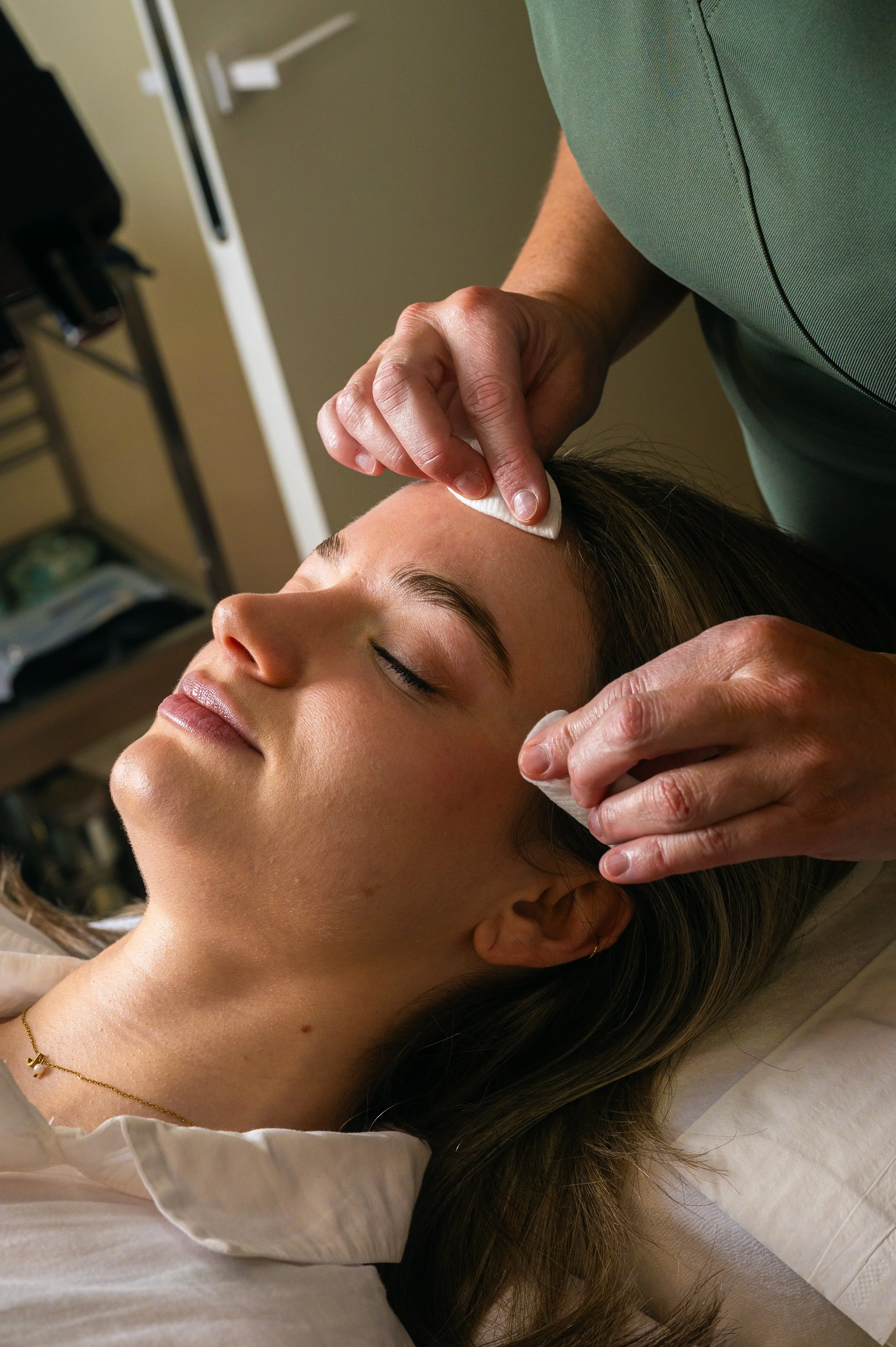 A woman receiving a facial treatment, with her eyes closed, while a professional gently wipes her face with cotton pads.